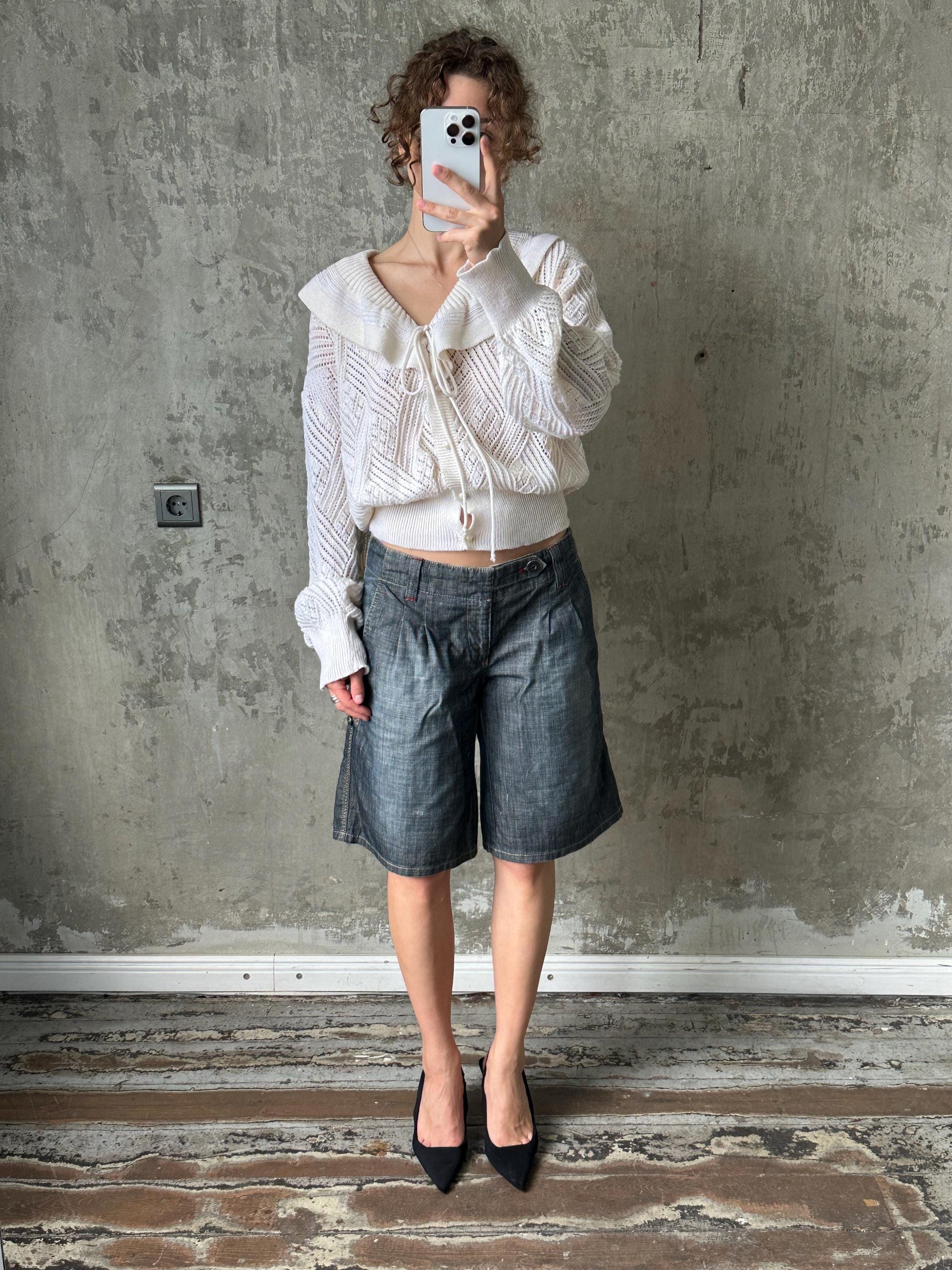 A person is taking a selfie with a white phone, standing in front of a concrete wall and wearing a white sweater, blue jeans, and black heels.