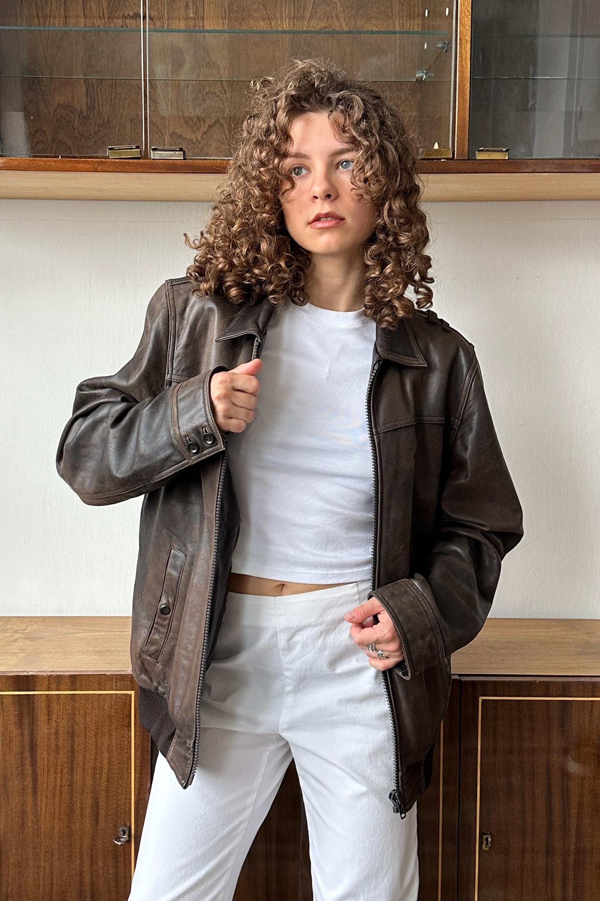 A woman with curly hair wearing a brown leather jacket and white pants stands in front of a wooden cabinet.