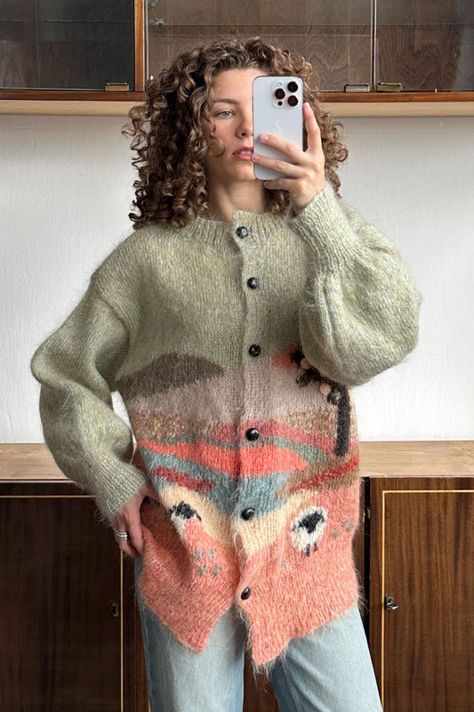 A woman with curly hair is taking a selfie with a smartphone, wearing a colorful, patterned cardigan.