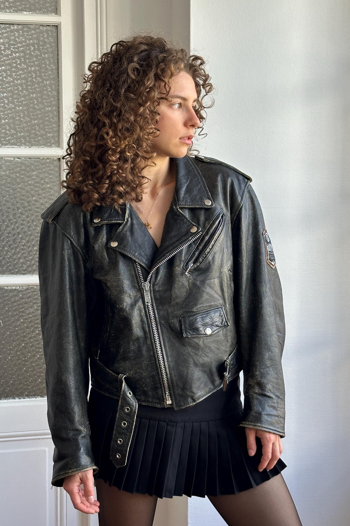 A woman with curly hair wearing a black leather jacket and a black pleated skirt stands in front of a white door.
