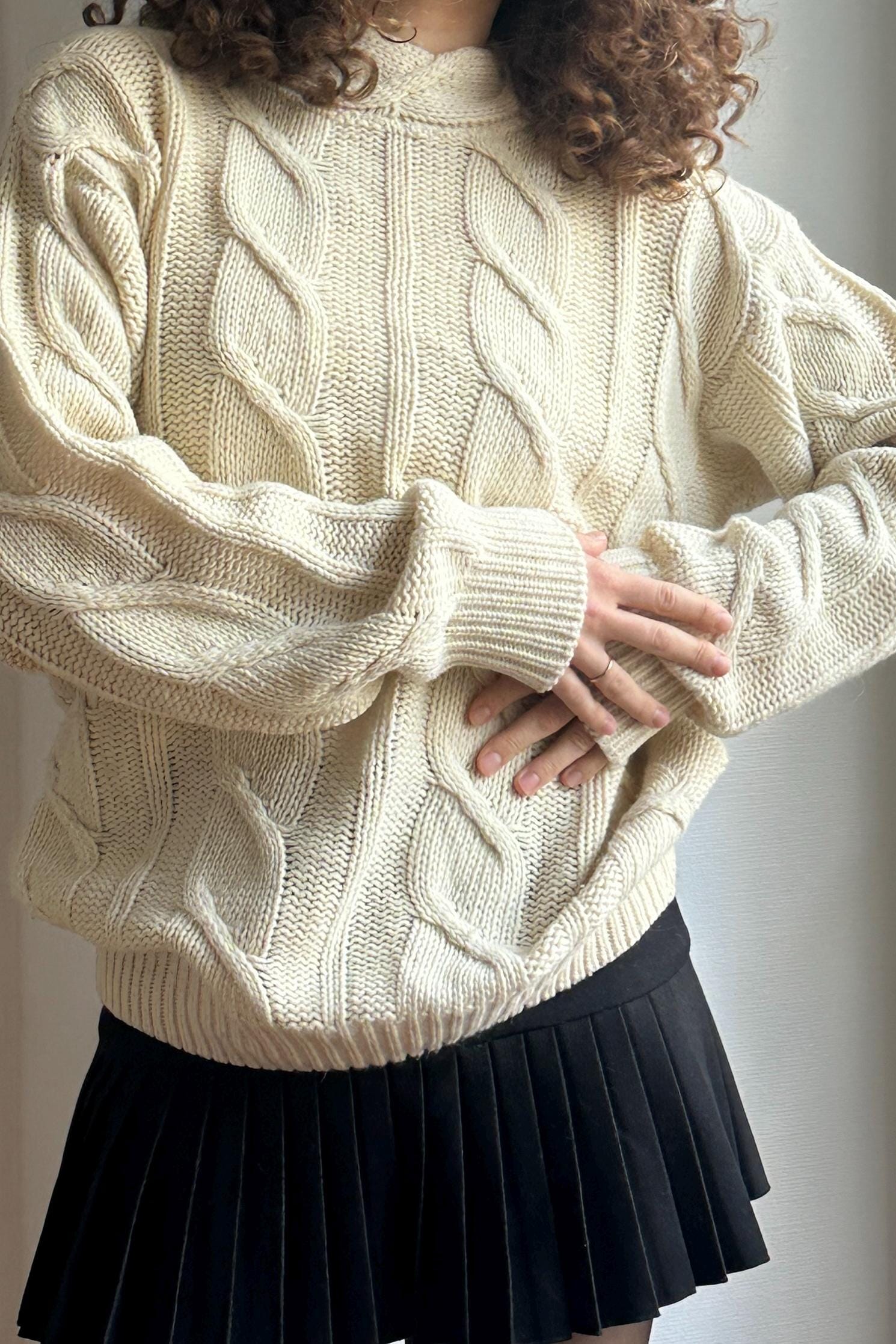 A person wearing a cream-colored knitted sweater and a black pleated skirt stands in front of a white wall.