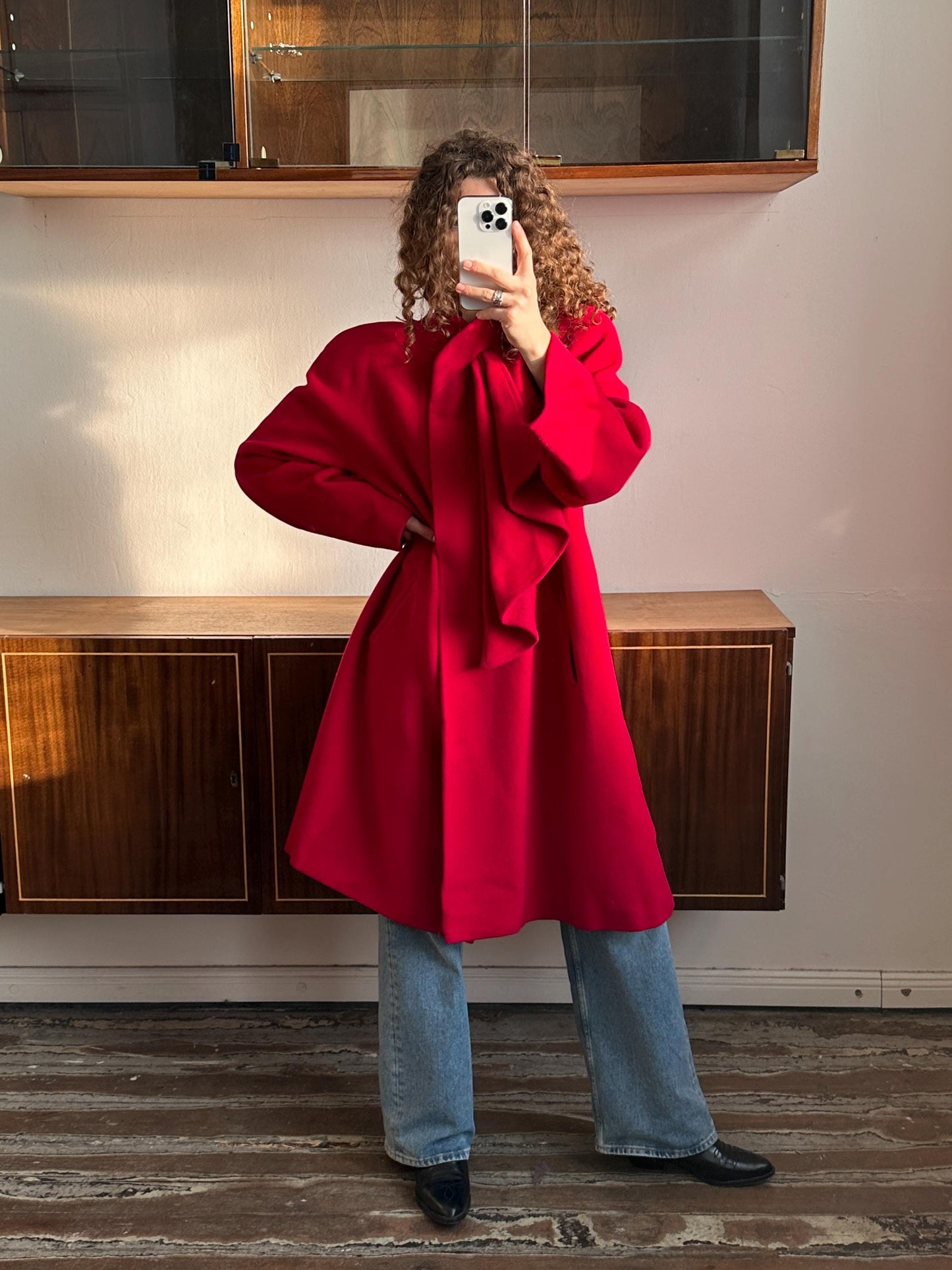 A person wearing a red coat is taking a selfie with a smartphone in front of a wooden cabinet.
