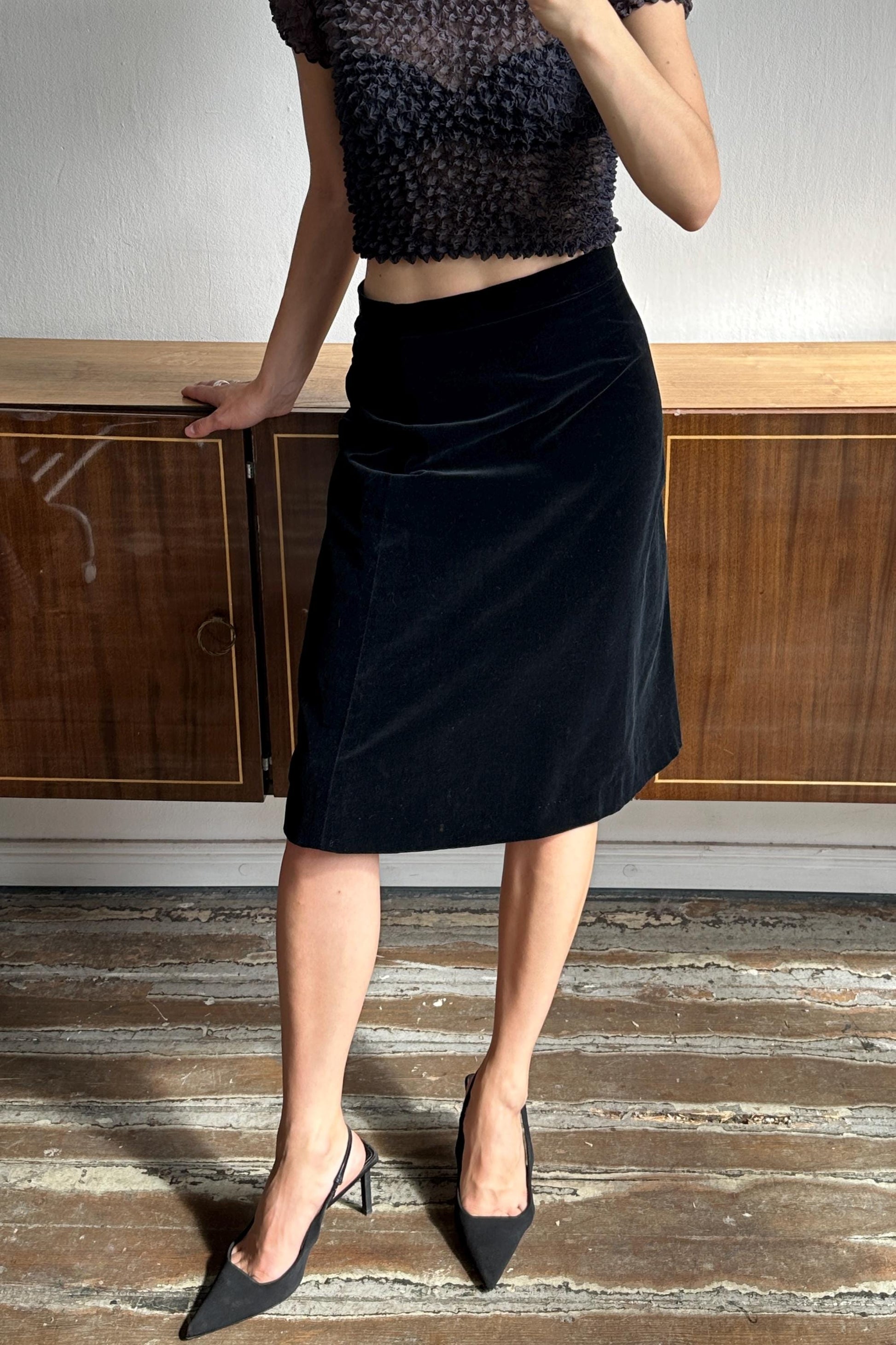 A woman wearing a black velvet skirt and a black top stands in front of a wooden cabinet.