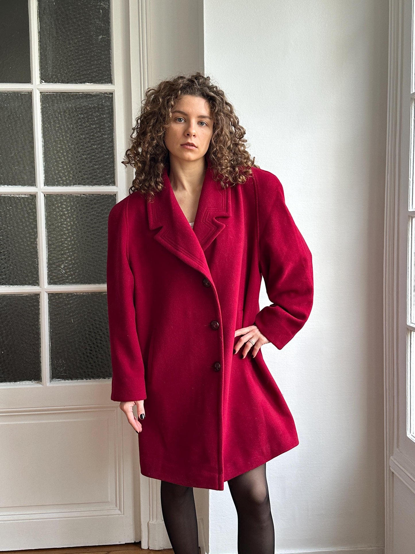 A woman with curly hair wearing a red coat is posing in a room with a white wall and a window.