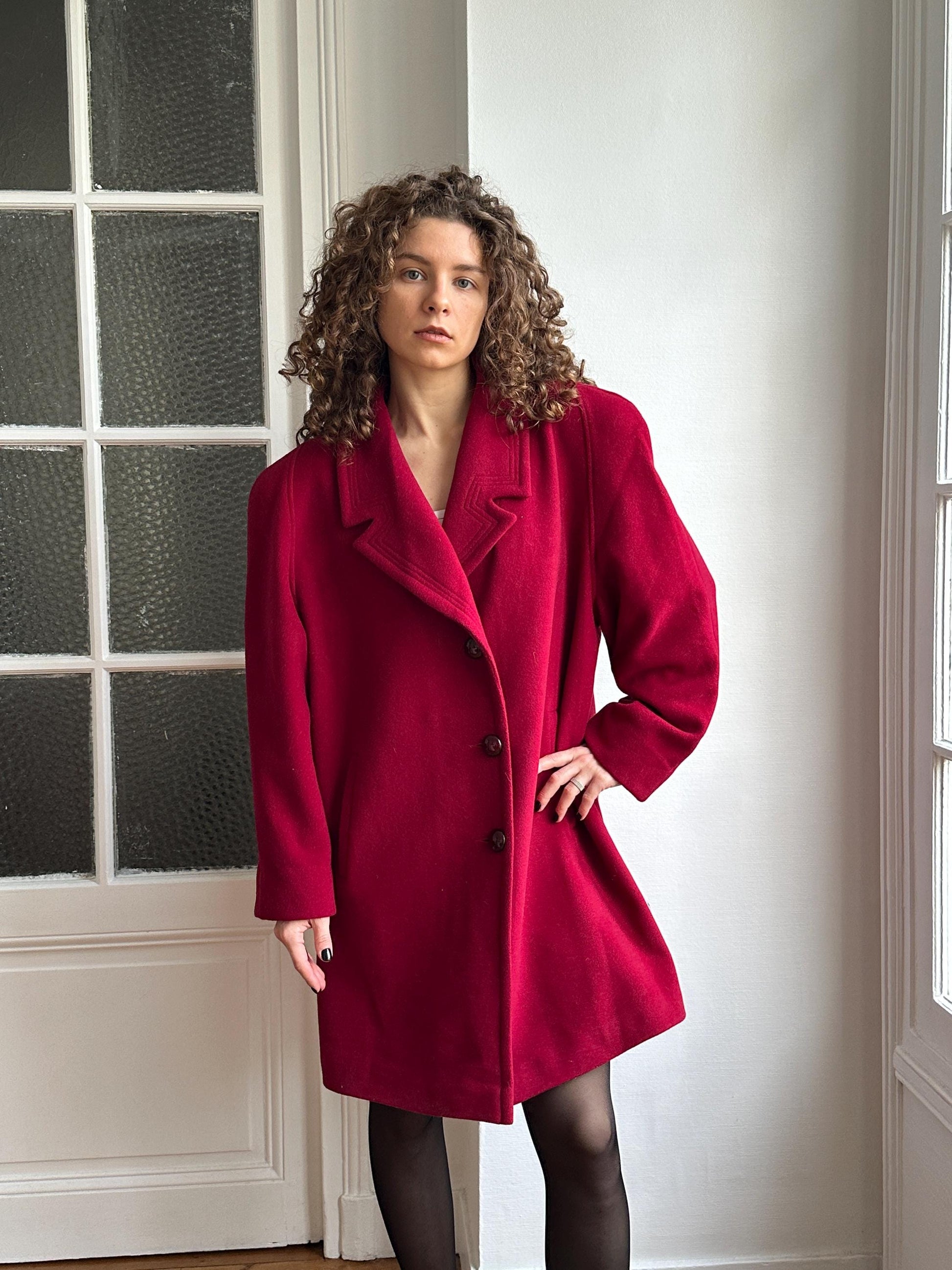 A woman with curly hair wearing a red coat is posing in a room with a white wall and a window.