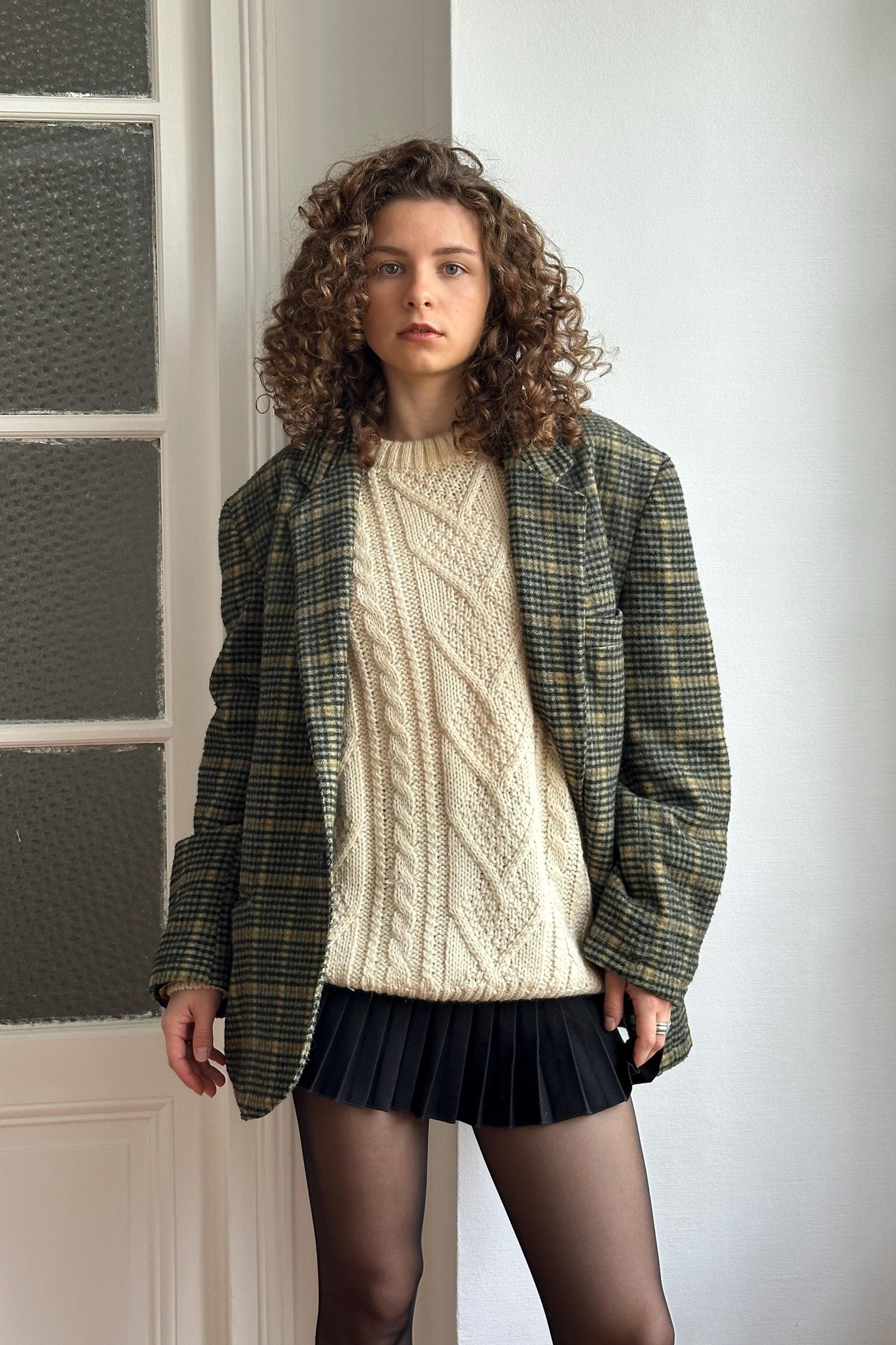 A young woman with curly hair stands in front of a white door, wearing a plaid jacket, a white sweater, and a black skirt.