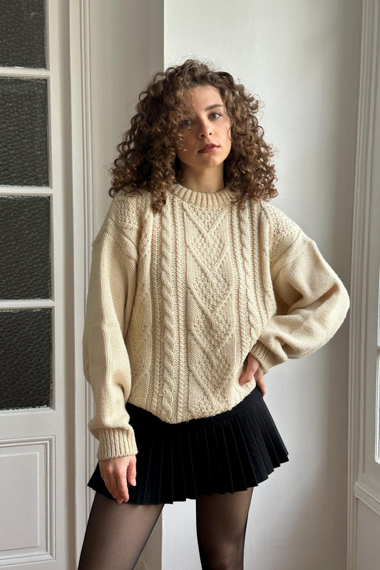 A young woman with curly hair stands in front of a white wall, wearing a cream-colored sweater and a black pleated skirt.