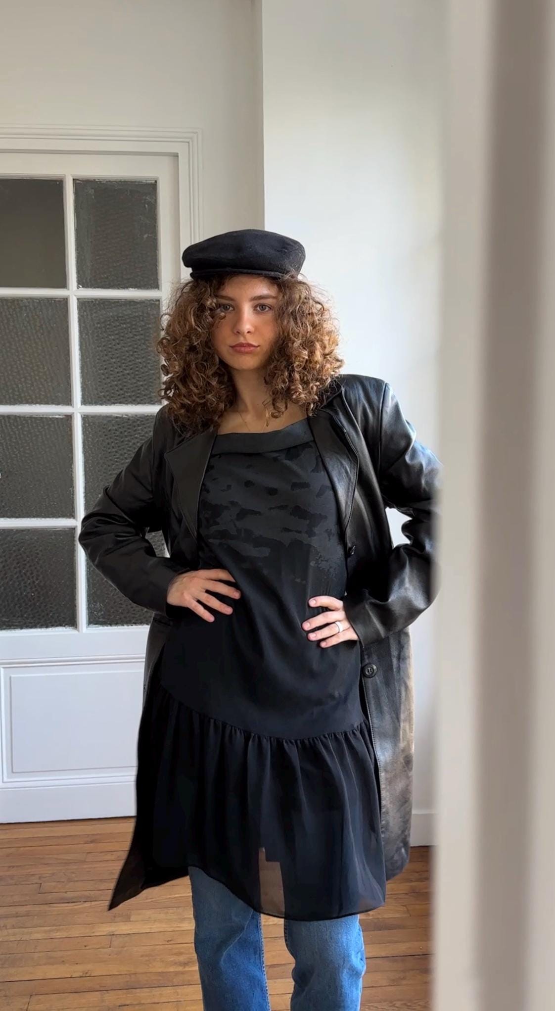 A woman with curly hair wearing a black coat, hat, and dress stands in a room with a white door and window.