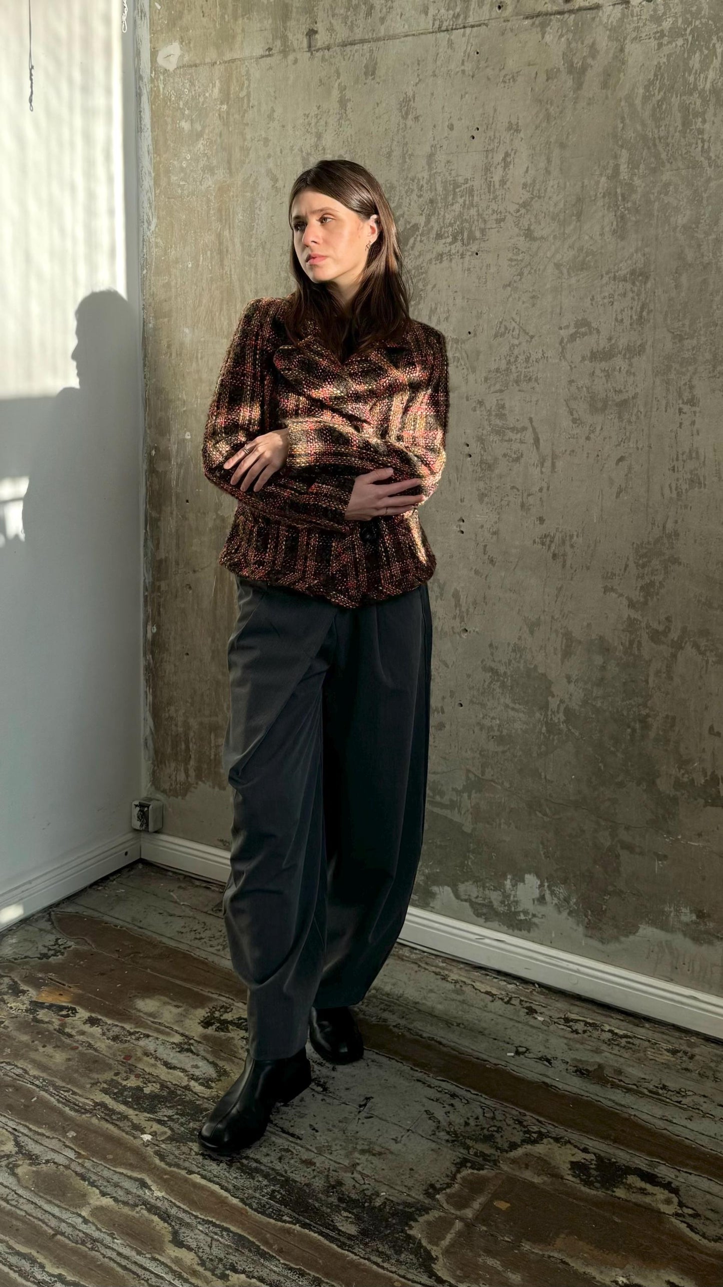A woman wearing a patterned jacket stands in a room with a concrete wall and wooden floor.