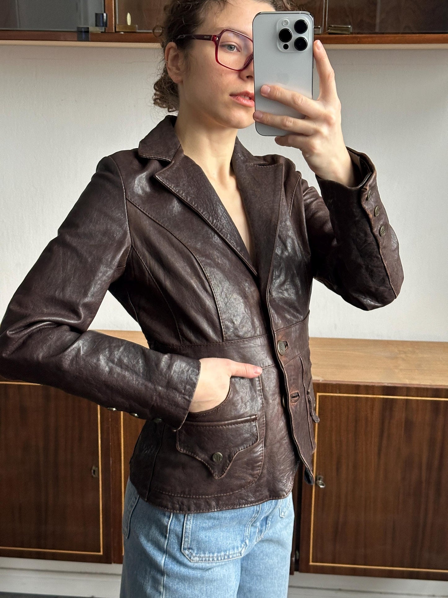 A person wearing a brown leather jacket and jeans is taking a selfie with a smartphone.