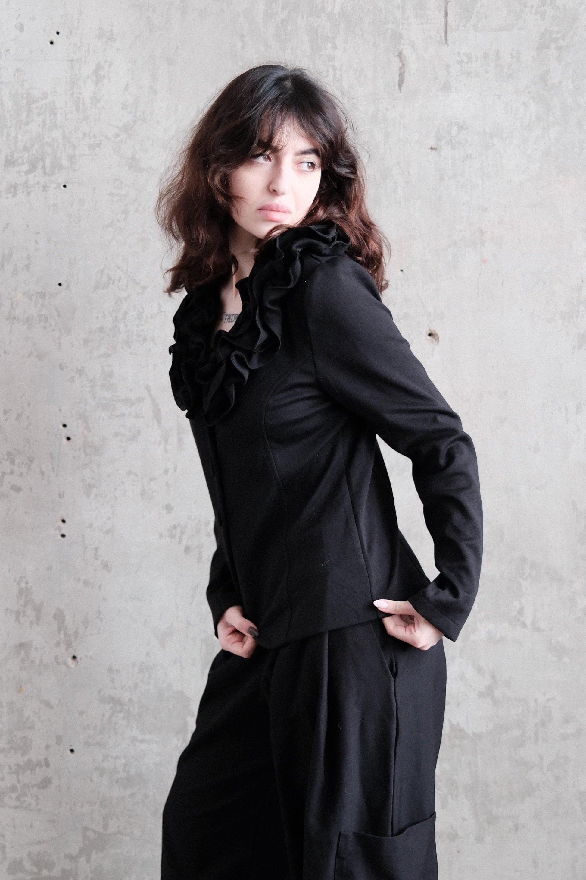 A woman in a black outfit poses against a concrete wall, her gaze directed off to the side.