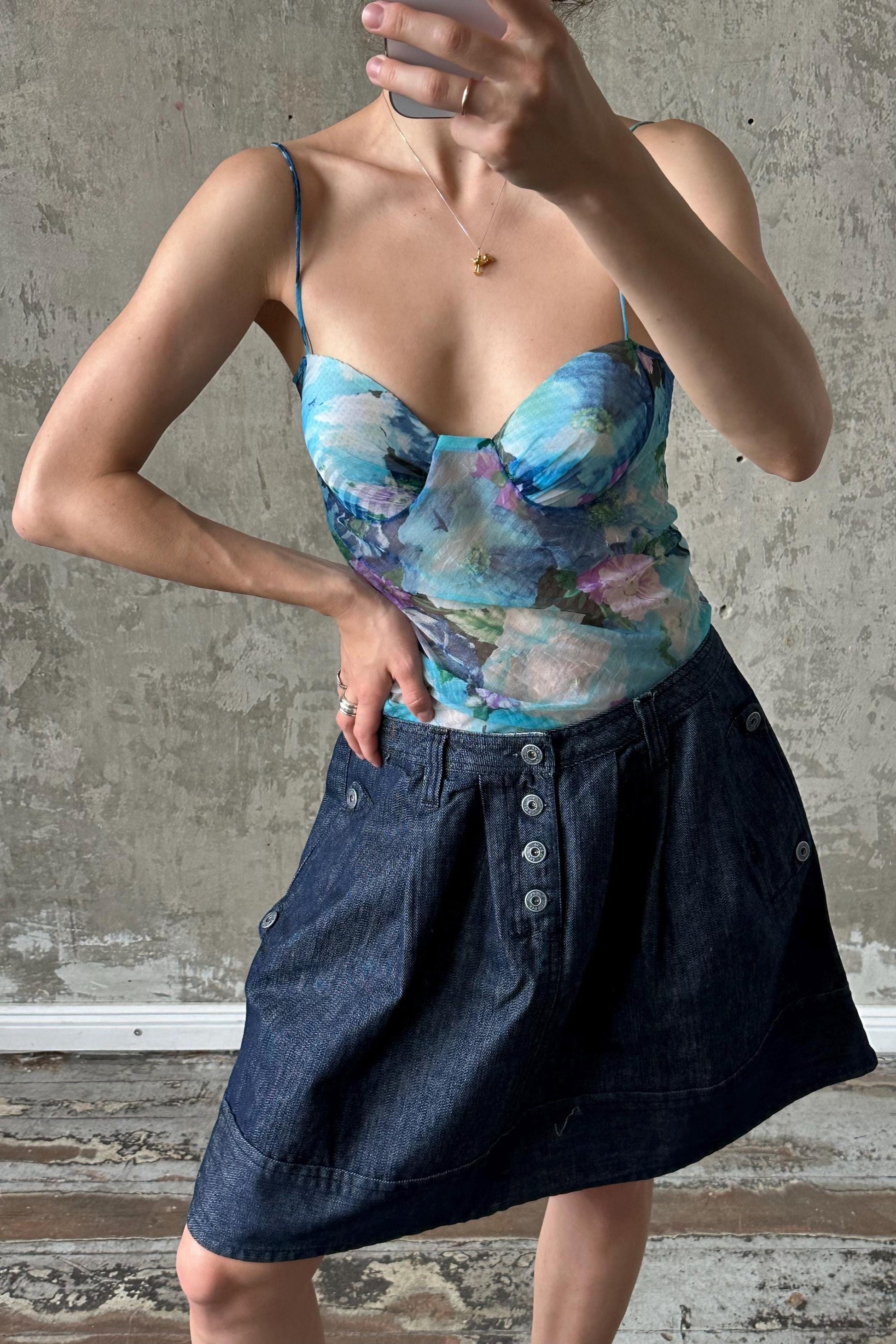 A woman is taking a selfie with a floral print top and a denim skirt, standing in front of a concrete wall.