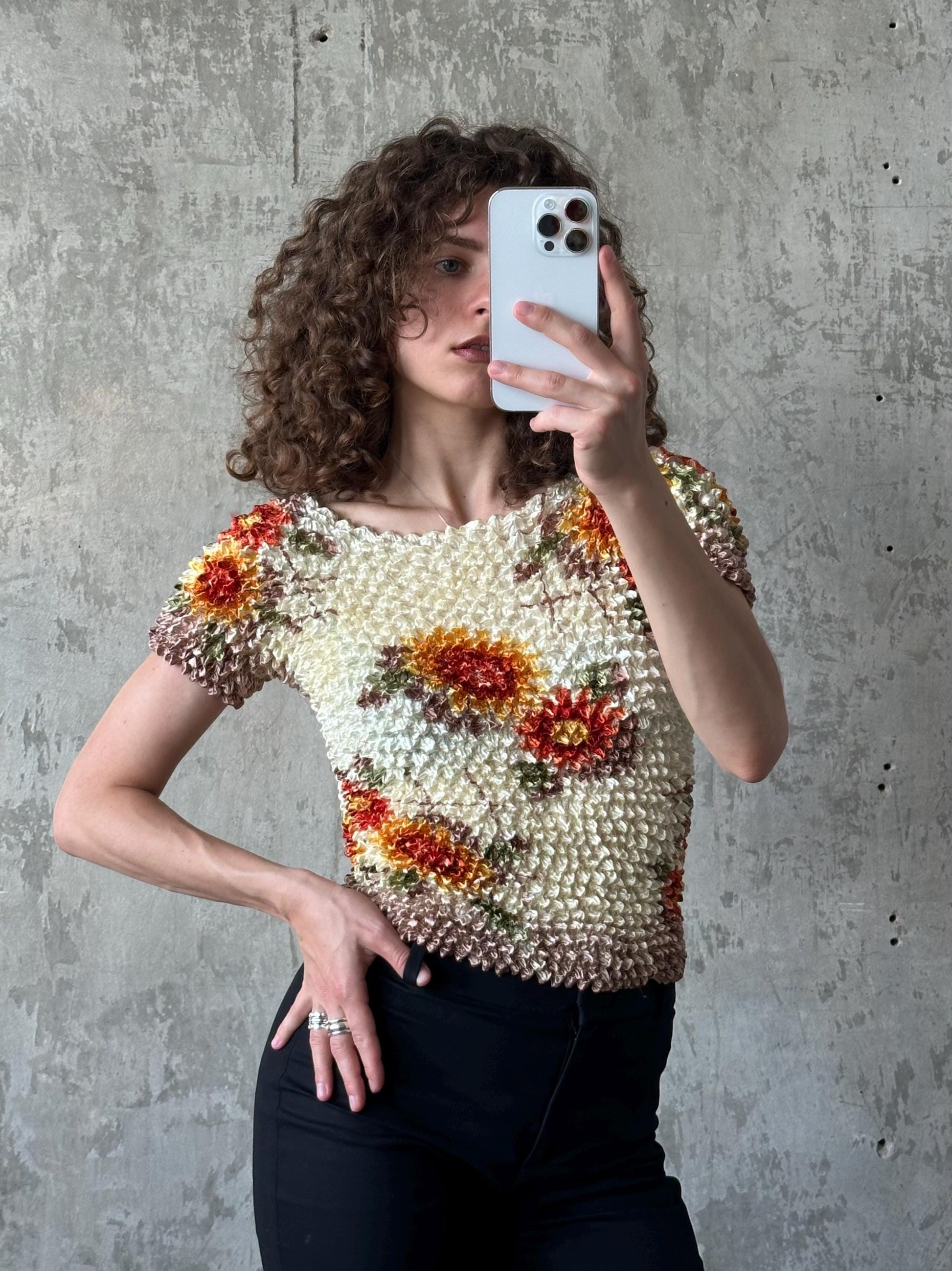 A woman with curly hair is taking a selfie with a white iPhone, wearing a floral print top and black pants.