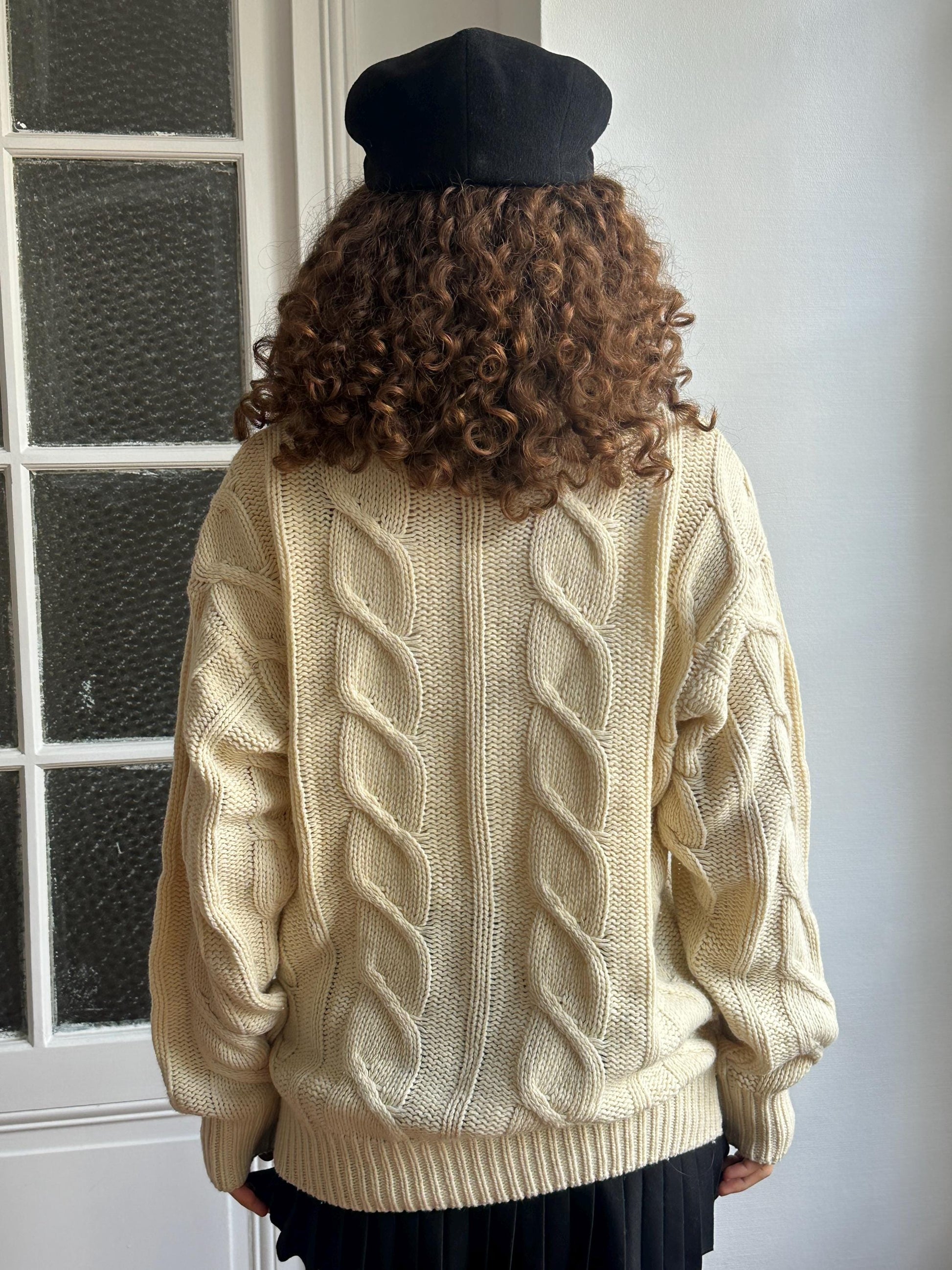 A person with curly hair wearing a cream-colored knitted sweater and a black hat stands in front of a window.