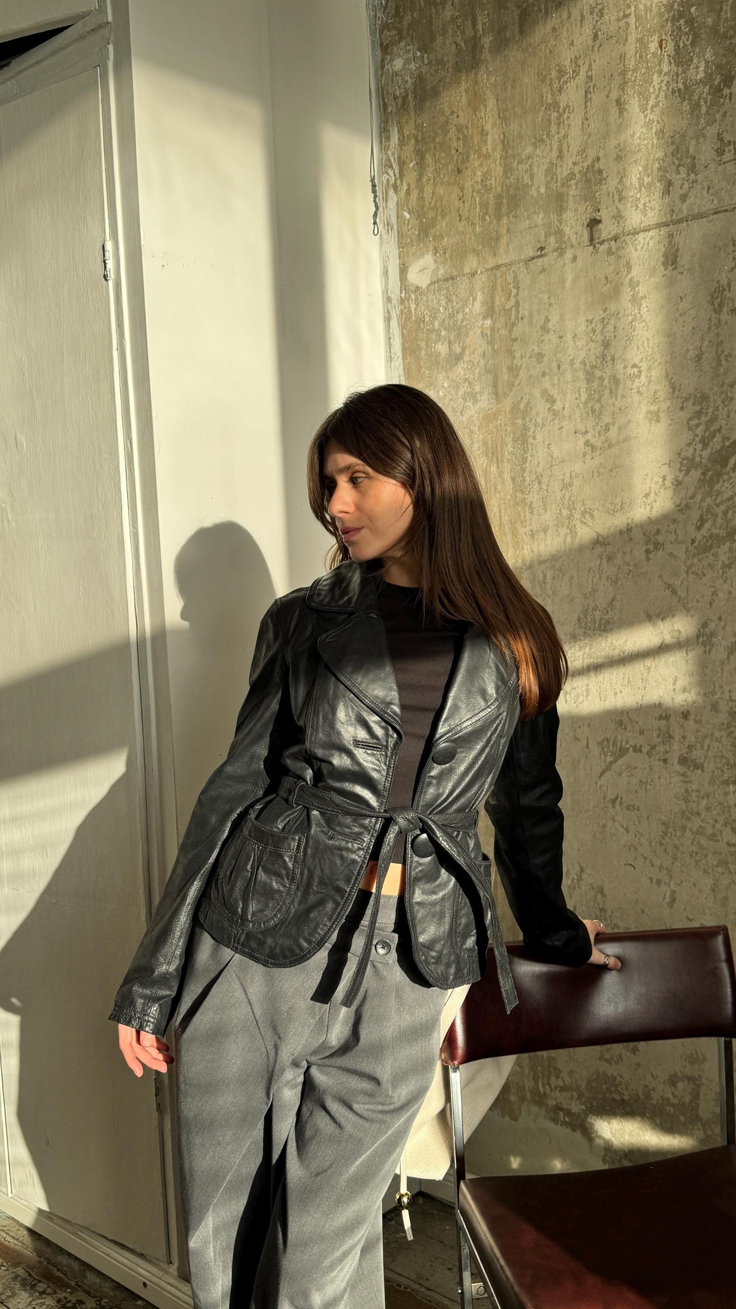 A woman in a black leather jacket stands in a room with a concrete wall and a white door, leaning against a chair.