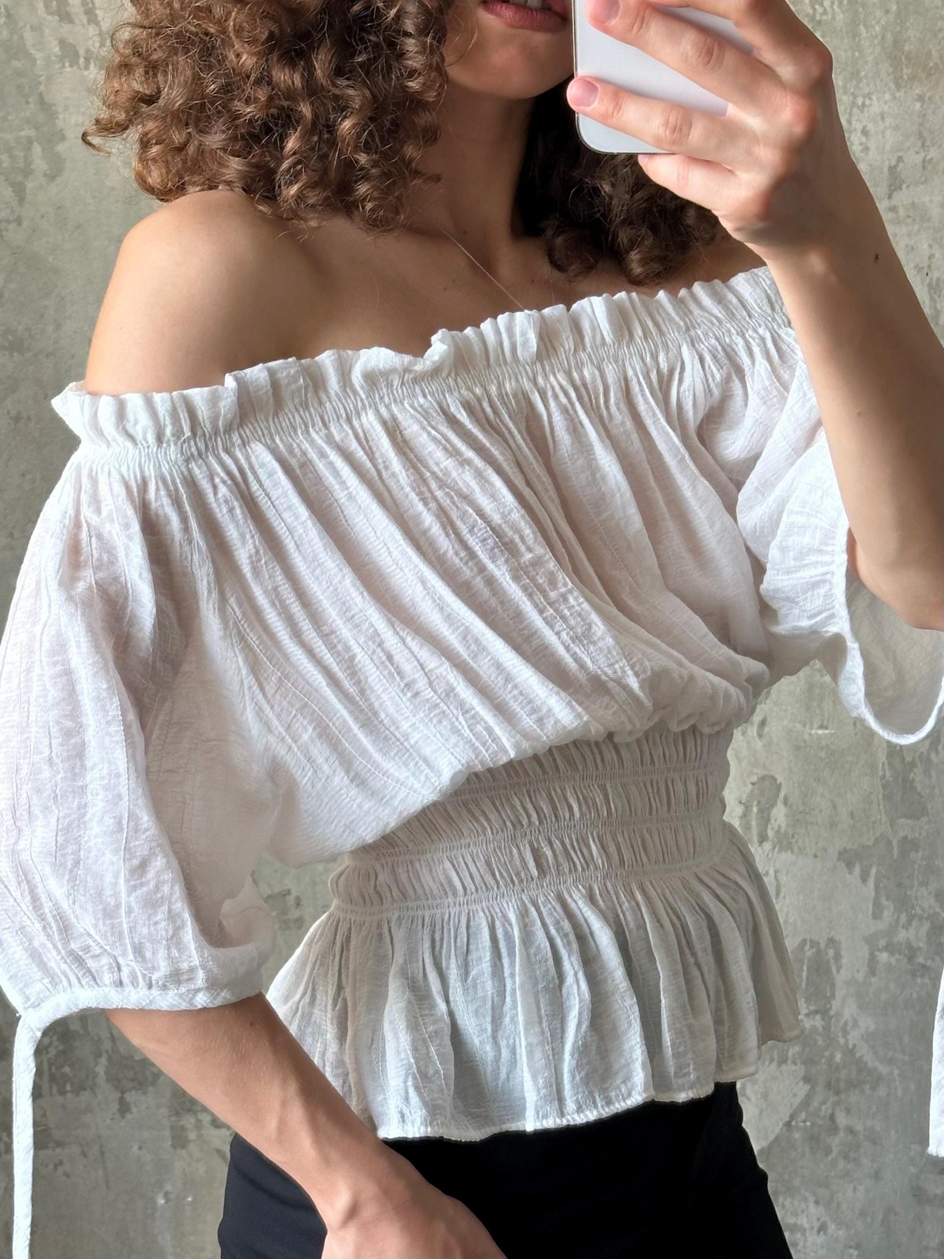 A woman with curly hair is taking a selfie with a white off-the-shoulder blouse, using a smartphone.