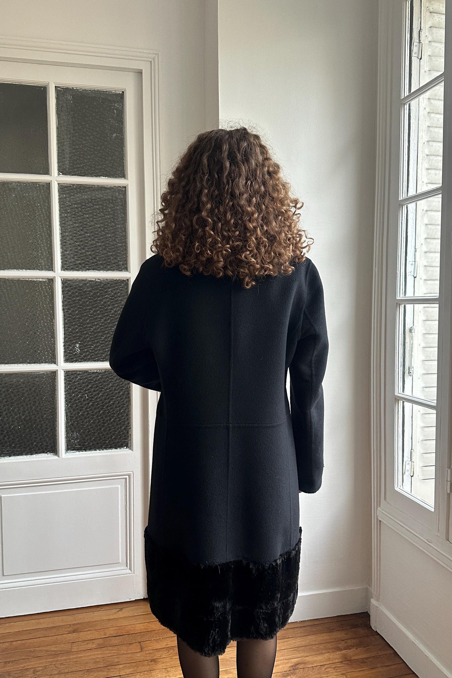 A person with curly hair wearing a black coat stands in a room with a window.
