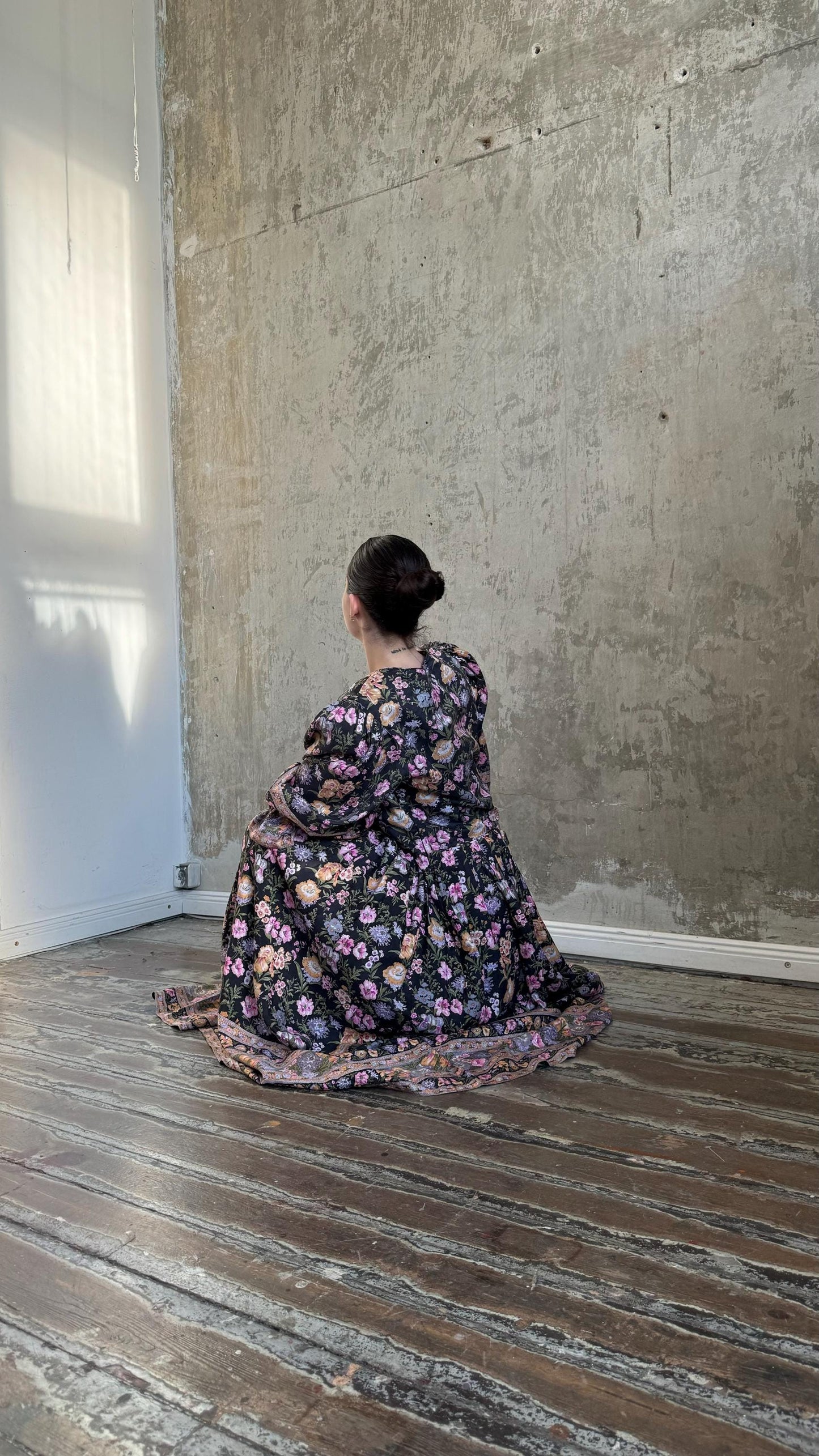 A person wearing a floral dress is sitting on a wooden floor in front of a concrete wall.