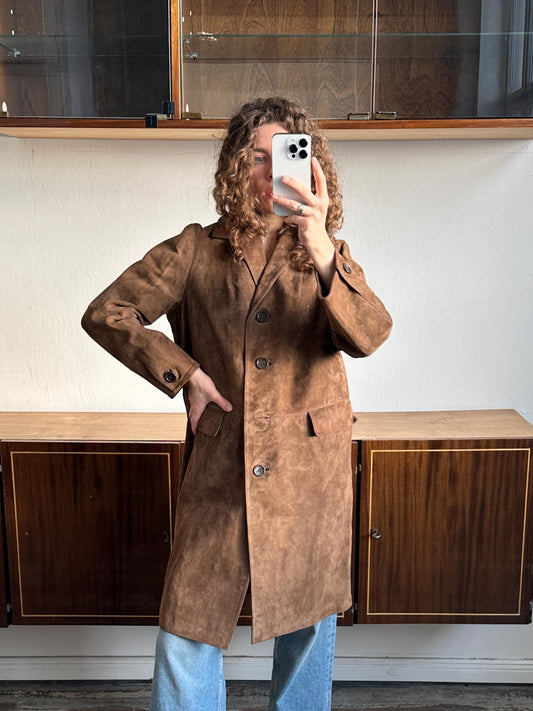 A person is taking a selfie with a brown suede coat, using a smartphone, in front of a wooden cabinet.