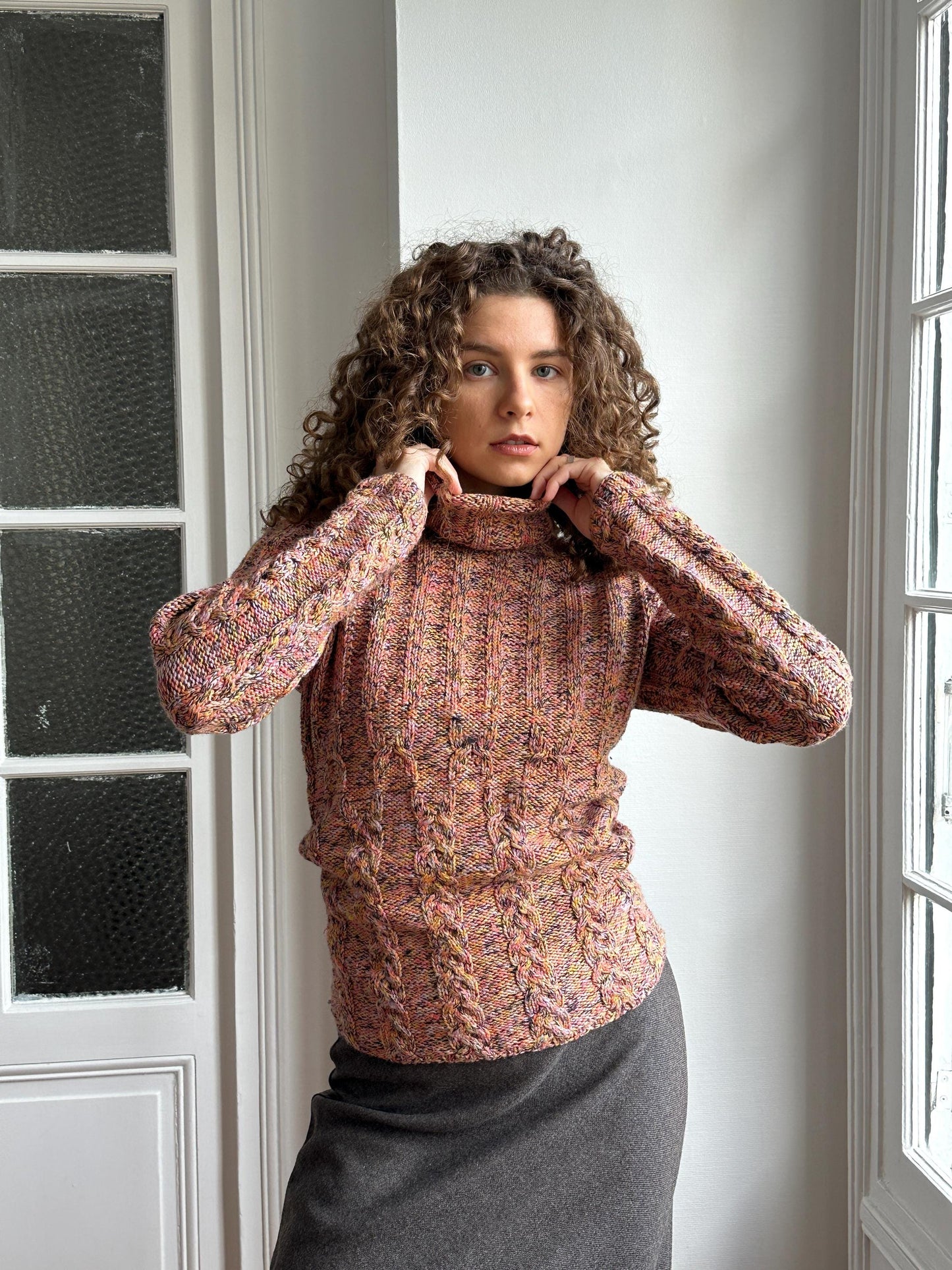 A woman with curly hair wearing a colorful sweater is posing in a room with a window.