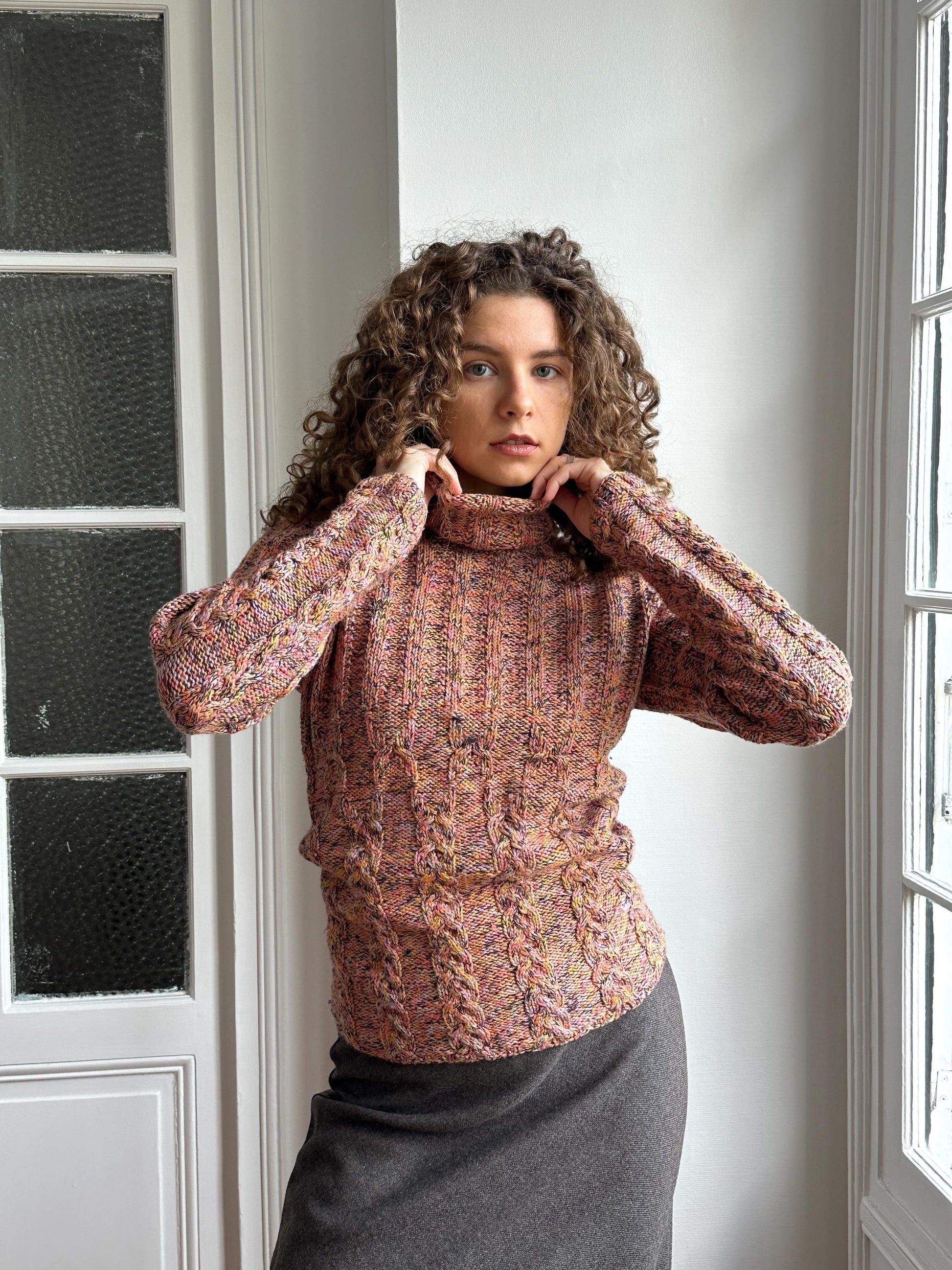 A woman with curly hair wearing a colorful sweater is posing in a room with a window.
