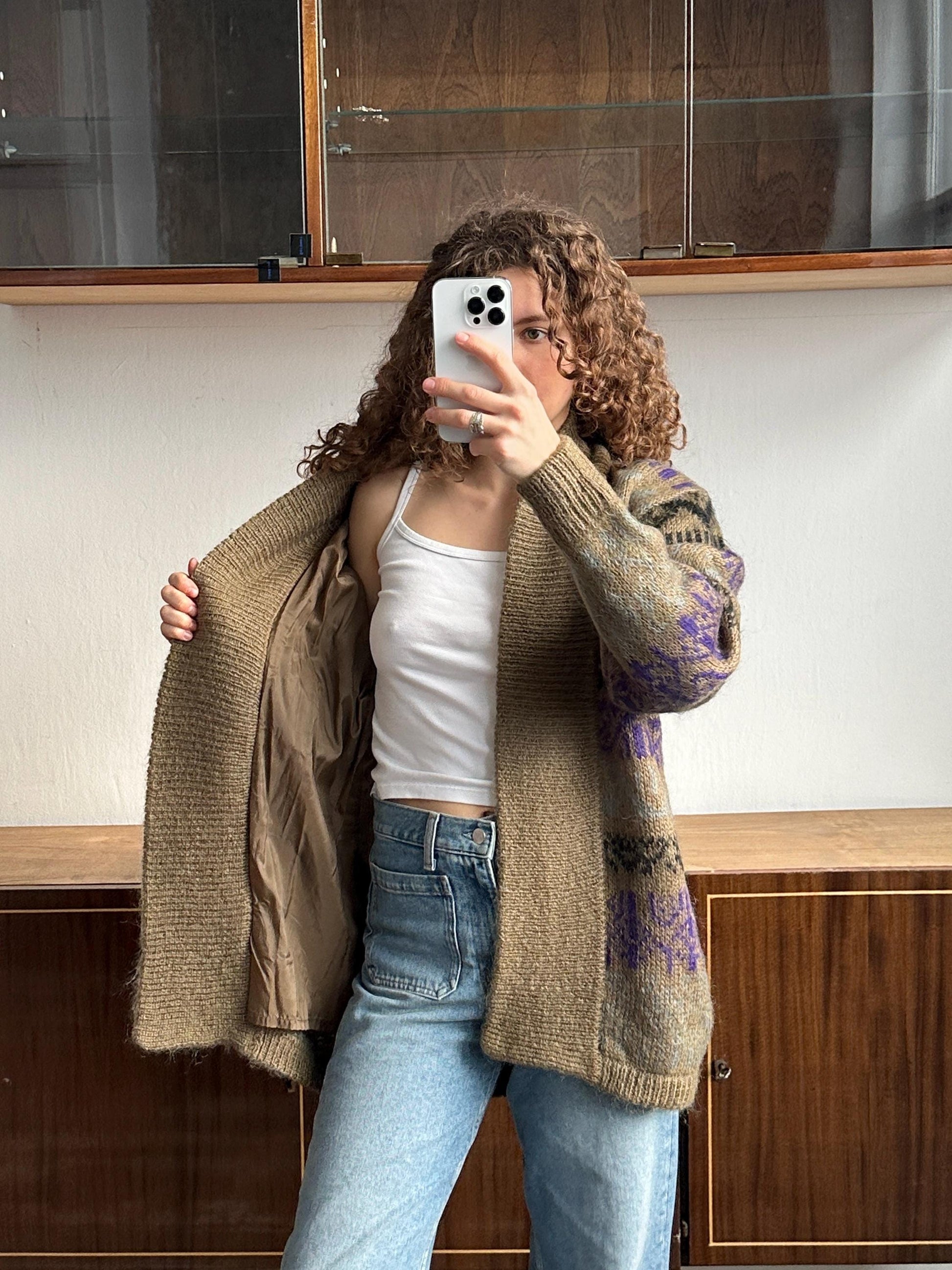 A woman with curly hair is taking a selfie with her phone, wearing a brown cardigan and a white tank top.