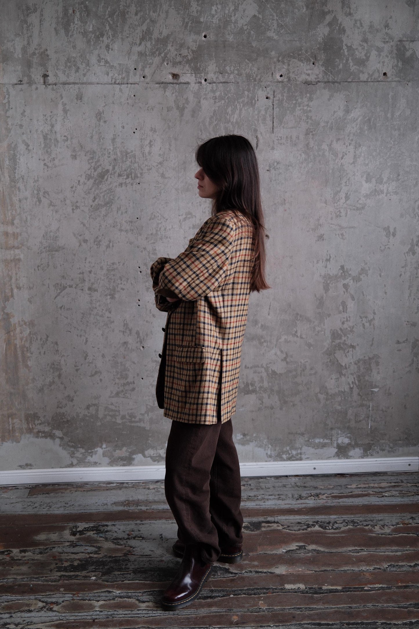 A person wearing a plaid jacket stands in a room with a concrete wall.