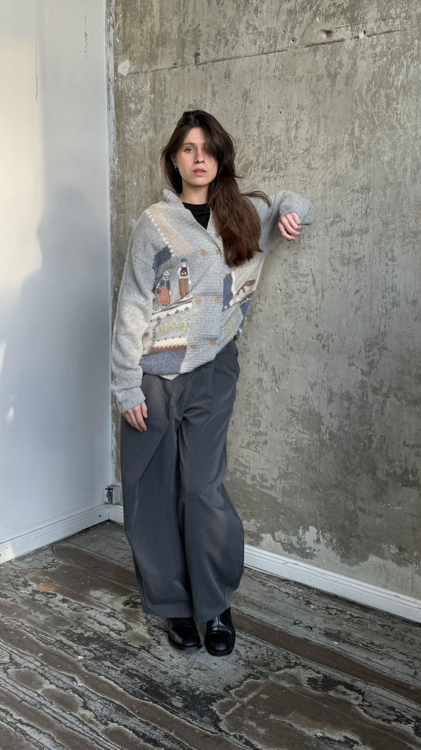 A woman wearing a gray sweater with a patterned design stands in a room with a concrete wall.
