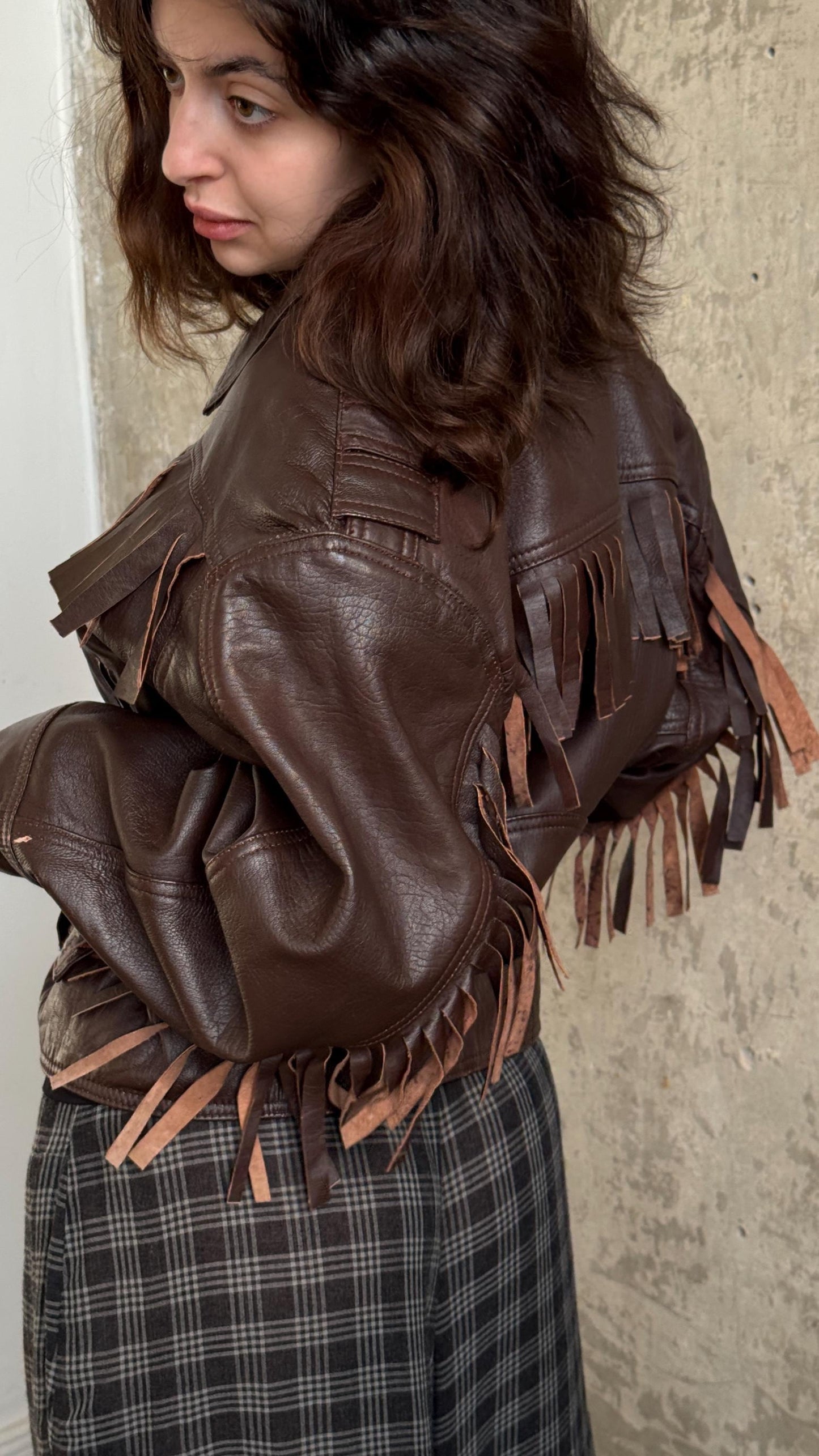 A woman wearing a brown leather jacket with fringe details stands against a concrete wall.