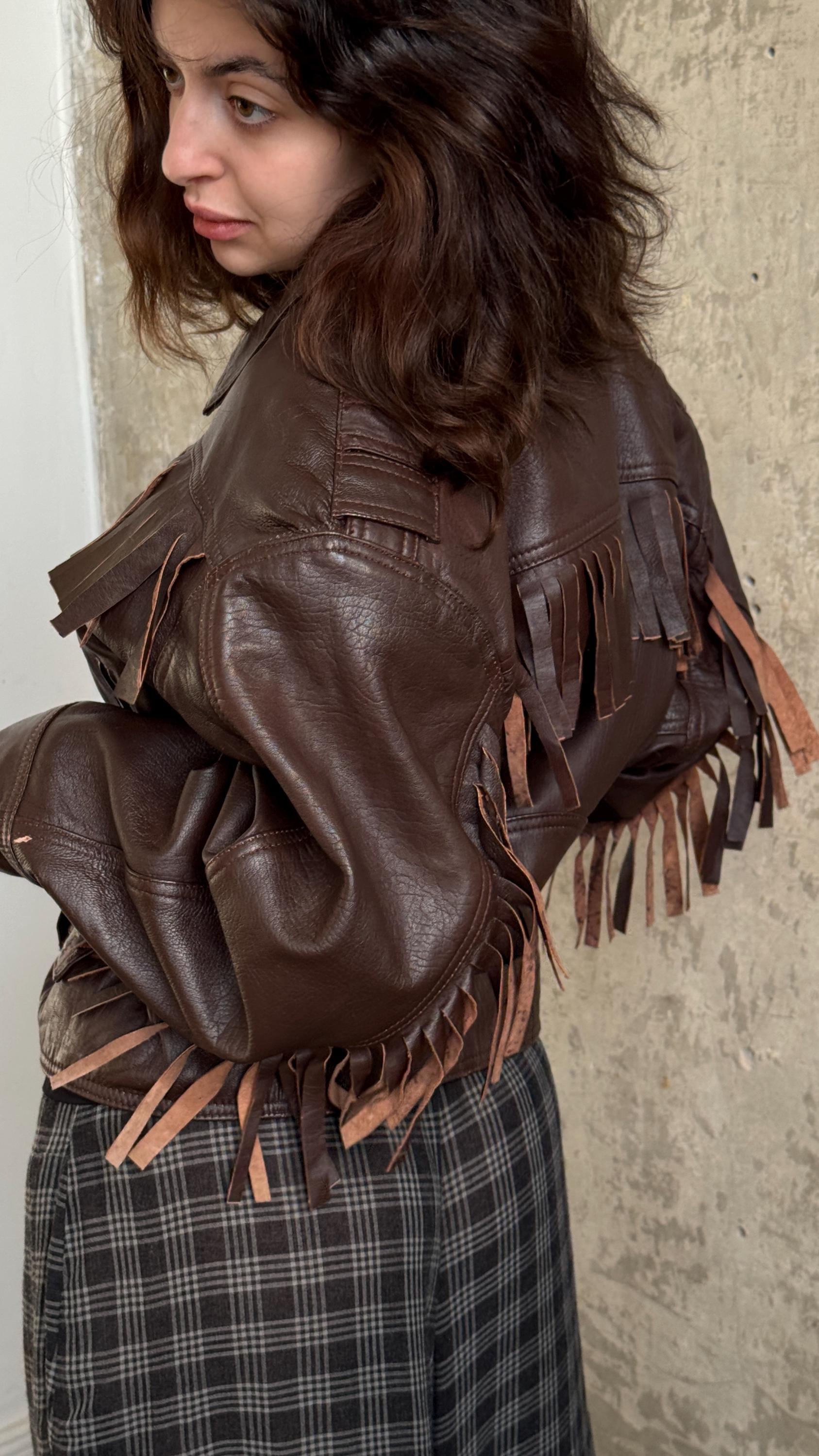 A woman wearing a brown leather jacket with fringe details stands against a concrete wall.