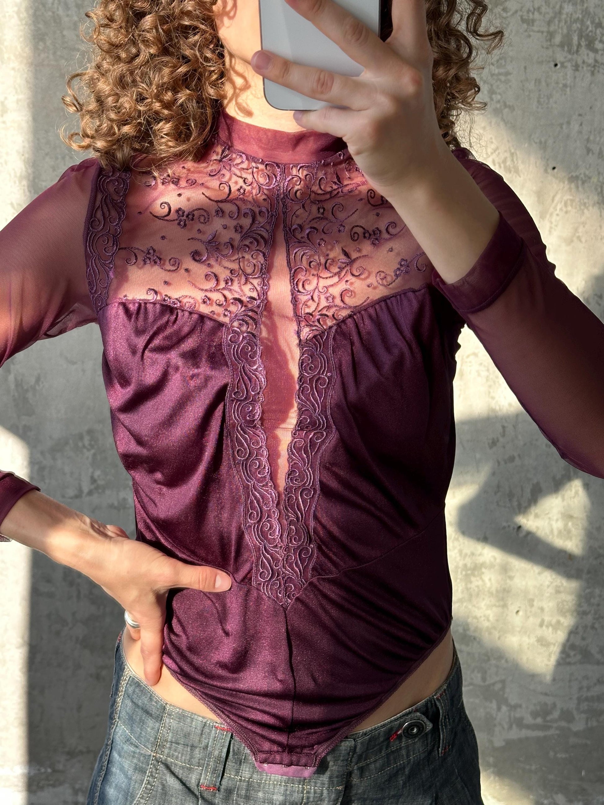 A person is taking a selfie with a smartphone, wearing a purple lace top and blue jeans.