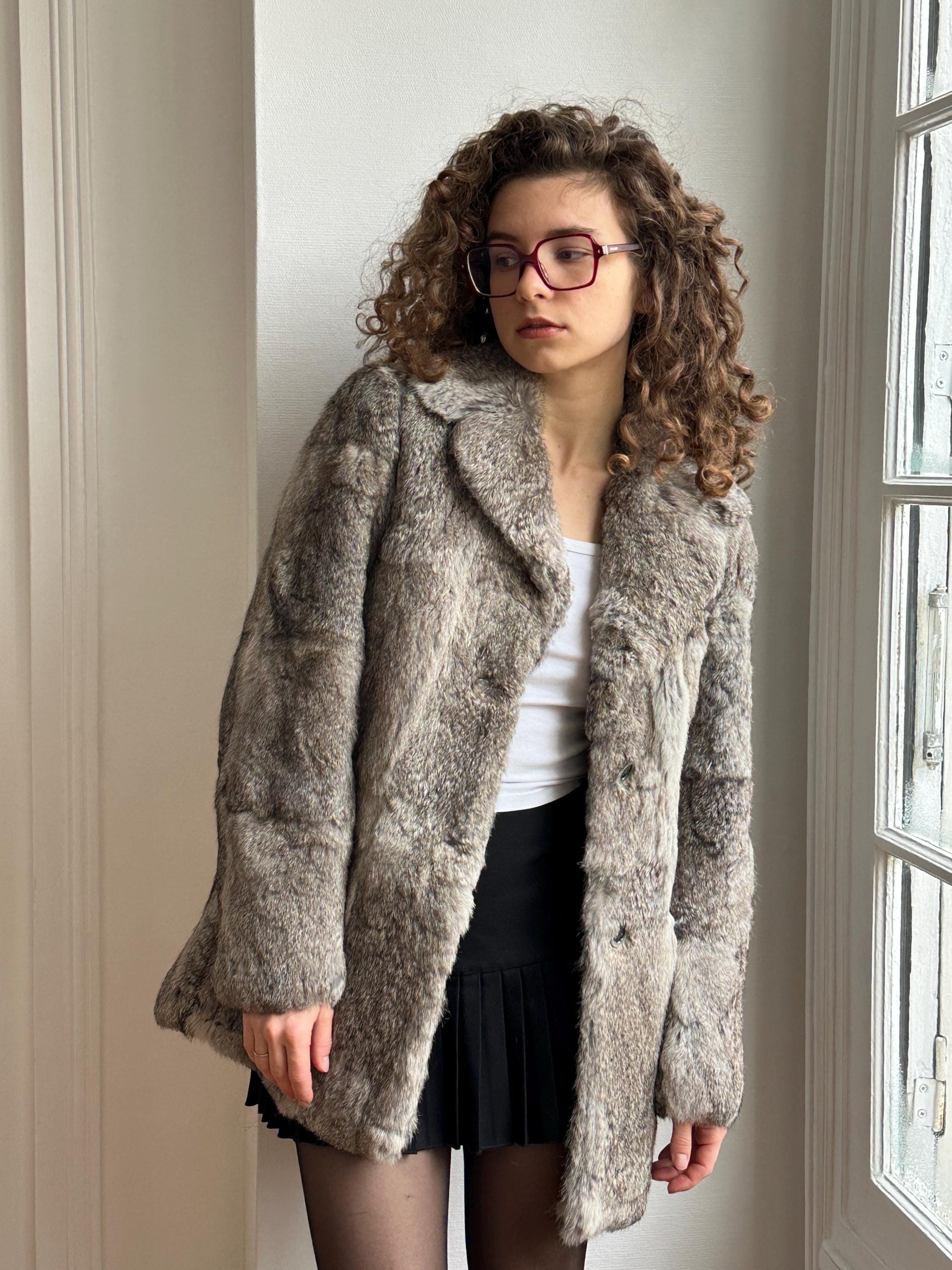 A young woman with curly hair and glasses stands in front of a window, wearing a gray fur coat and a black skirt.