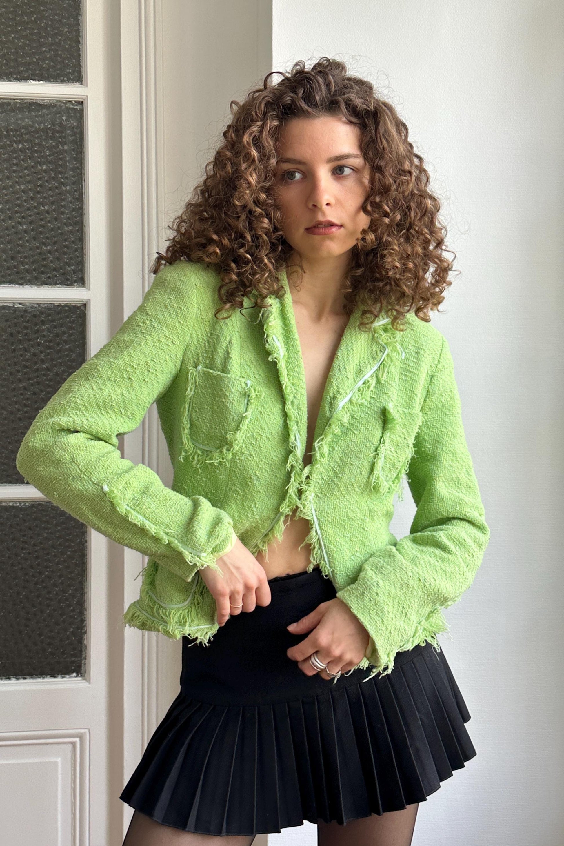 A woman with curly hair wearing a lime green tweed jacket and a black pleated skirt stands in front of a white door.