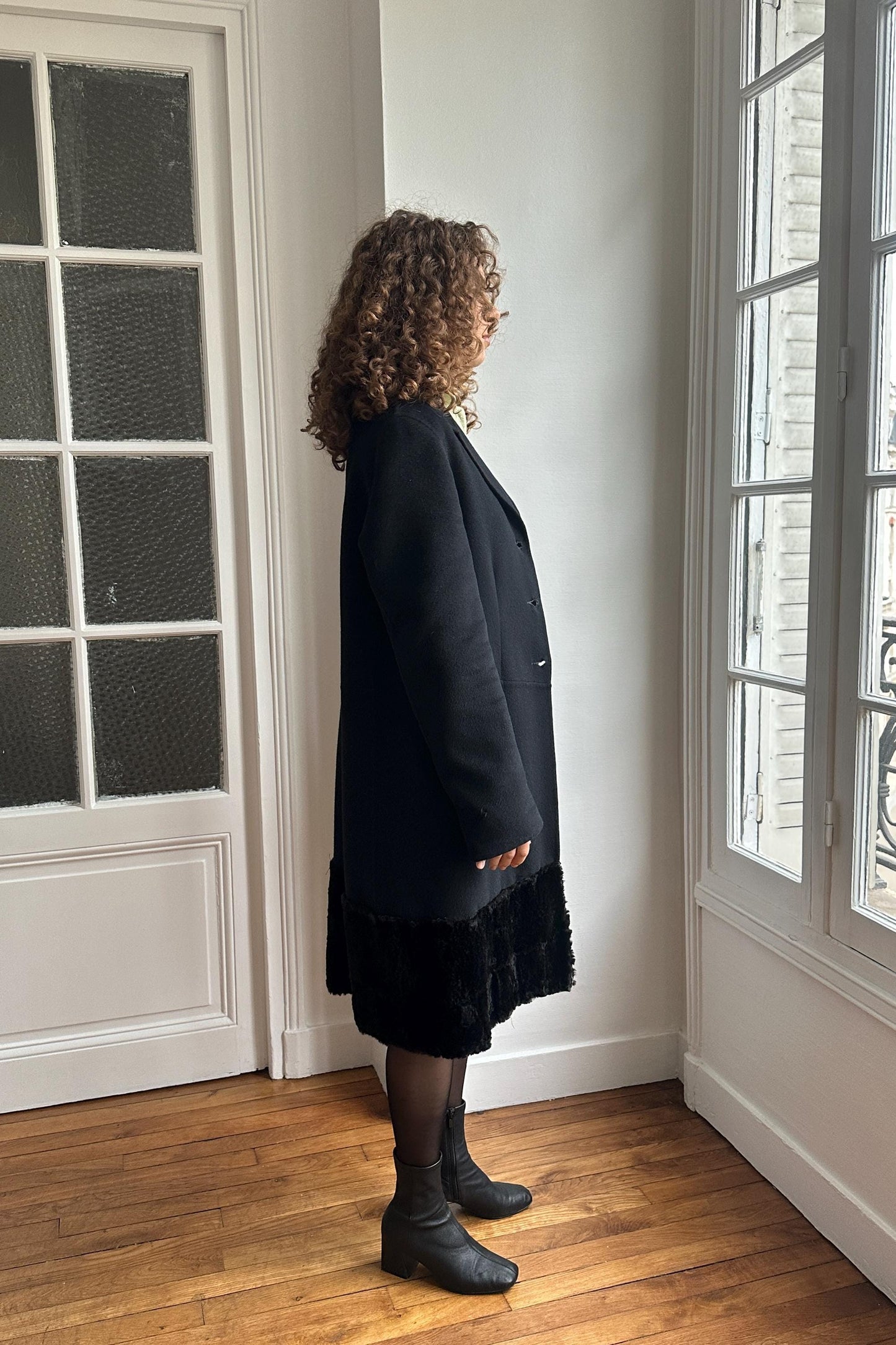 A woman with curly hair stands in a room, wearing a black coat and boots, facing away from the camera.