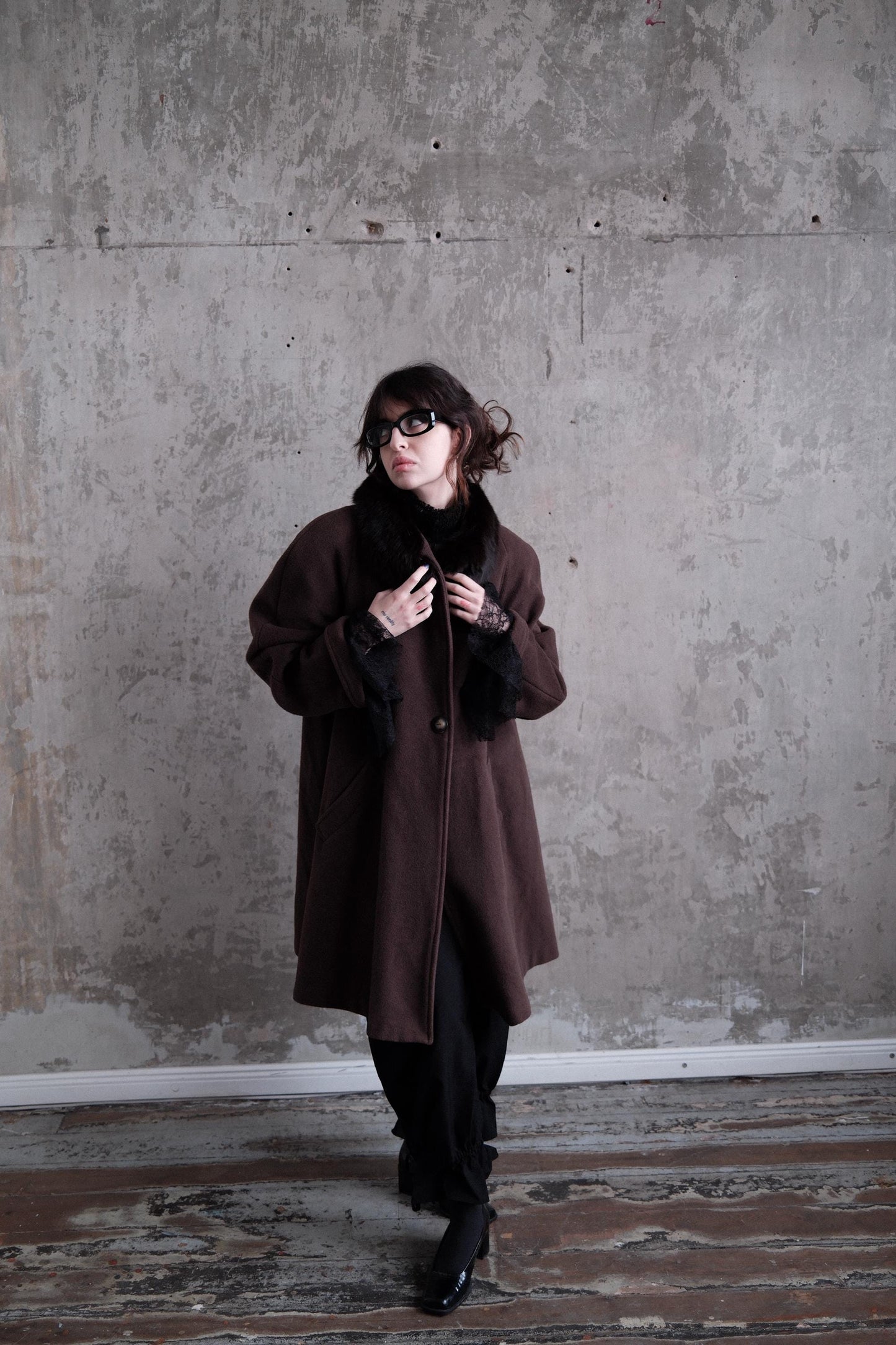 A woman in a brown coat stands in a room with a concrete wall, looking to her left.