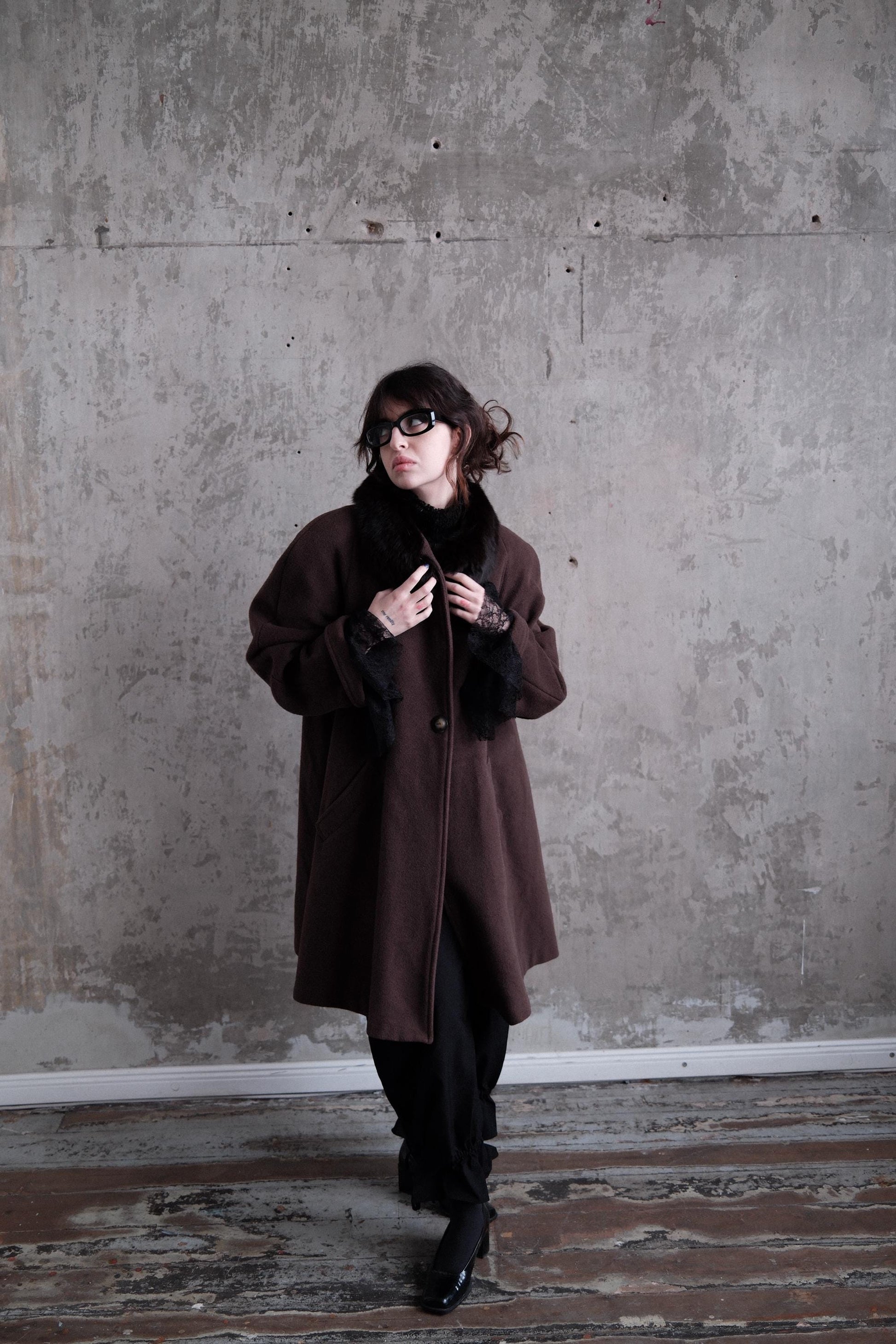 A woman in a brown coat stands in a room with a concrete wall, looking to her left.