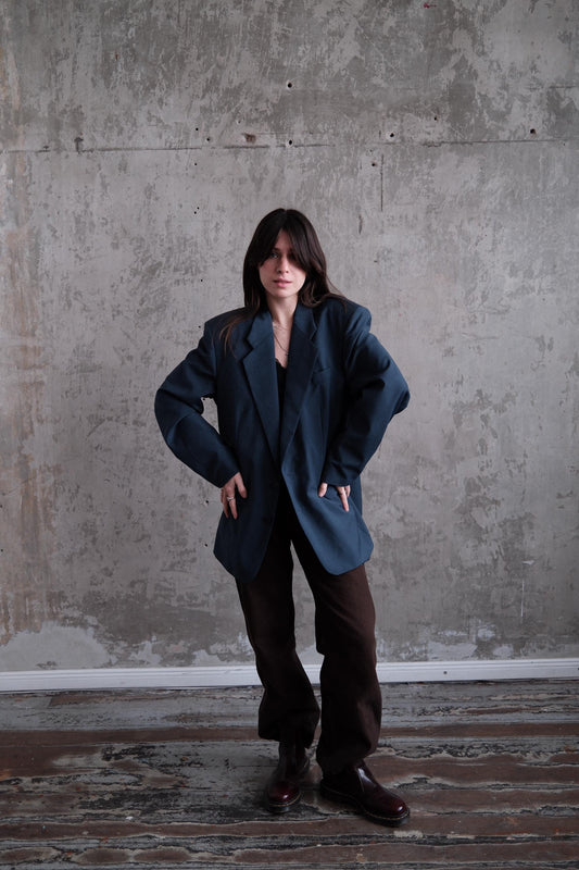 A person wearing a dark blue blazer and brown pants stands in a room with a concrete wall.