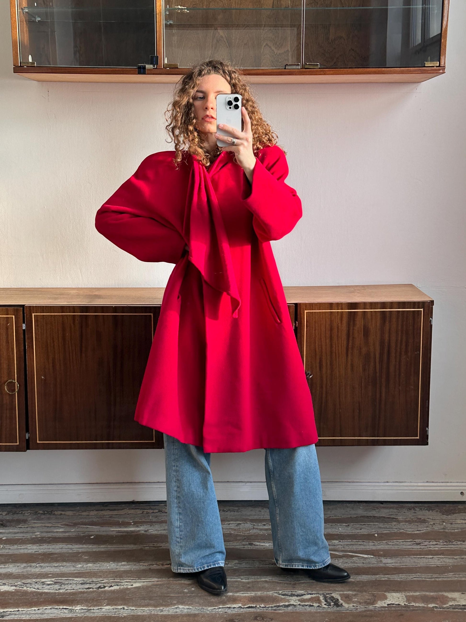 A woman in a red coat is taking a selfie with a white phone in front of a wooden cabinet.