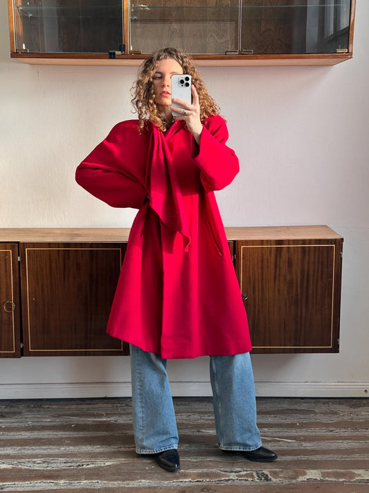 A woman in a red coat is taking a selfie with a white phone in front of a wooden cabinet.