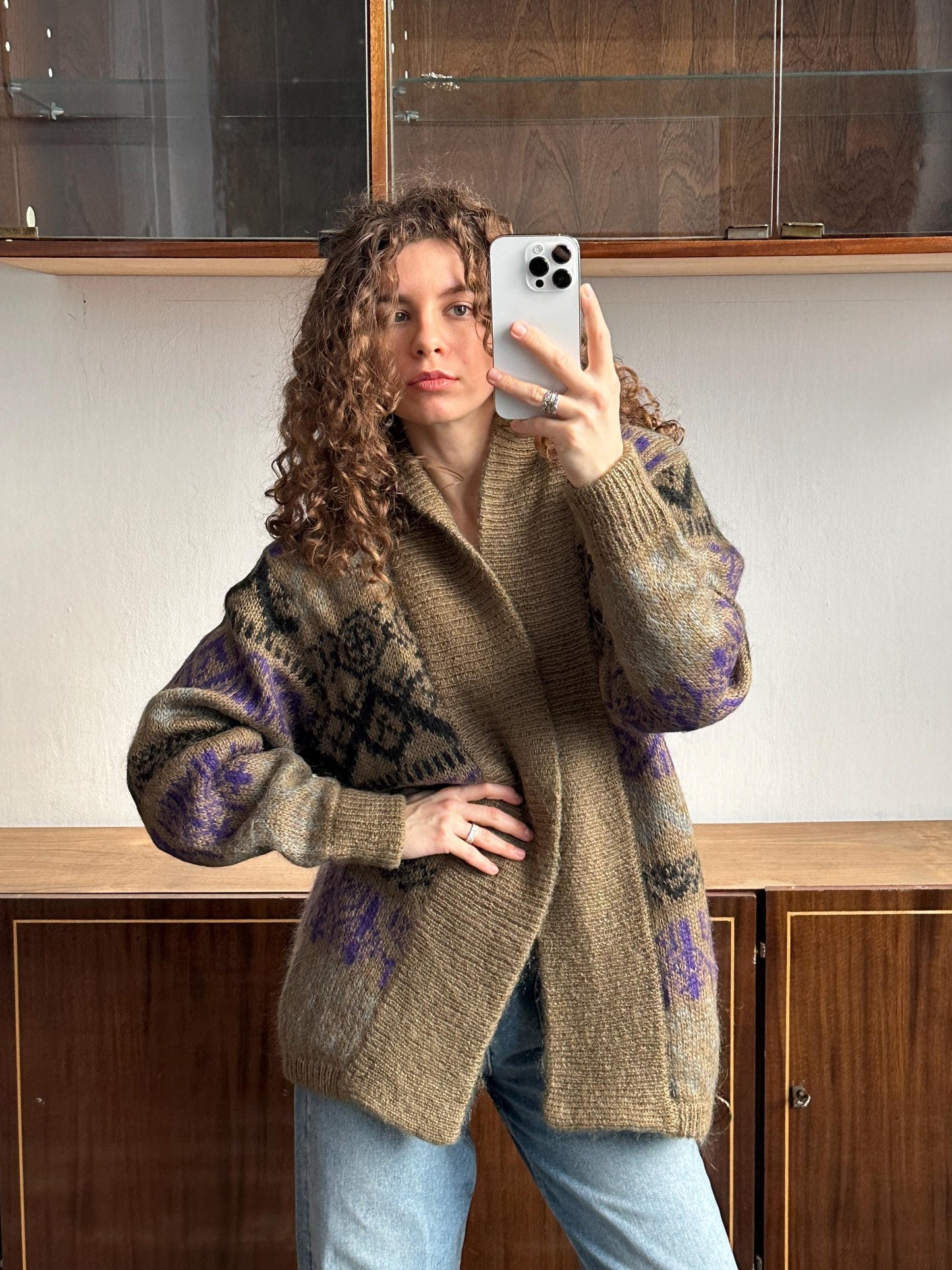 A woman with curly hair is taking a selfie with a white iPhone, wearing a patterned sweater and jeans.