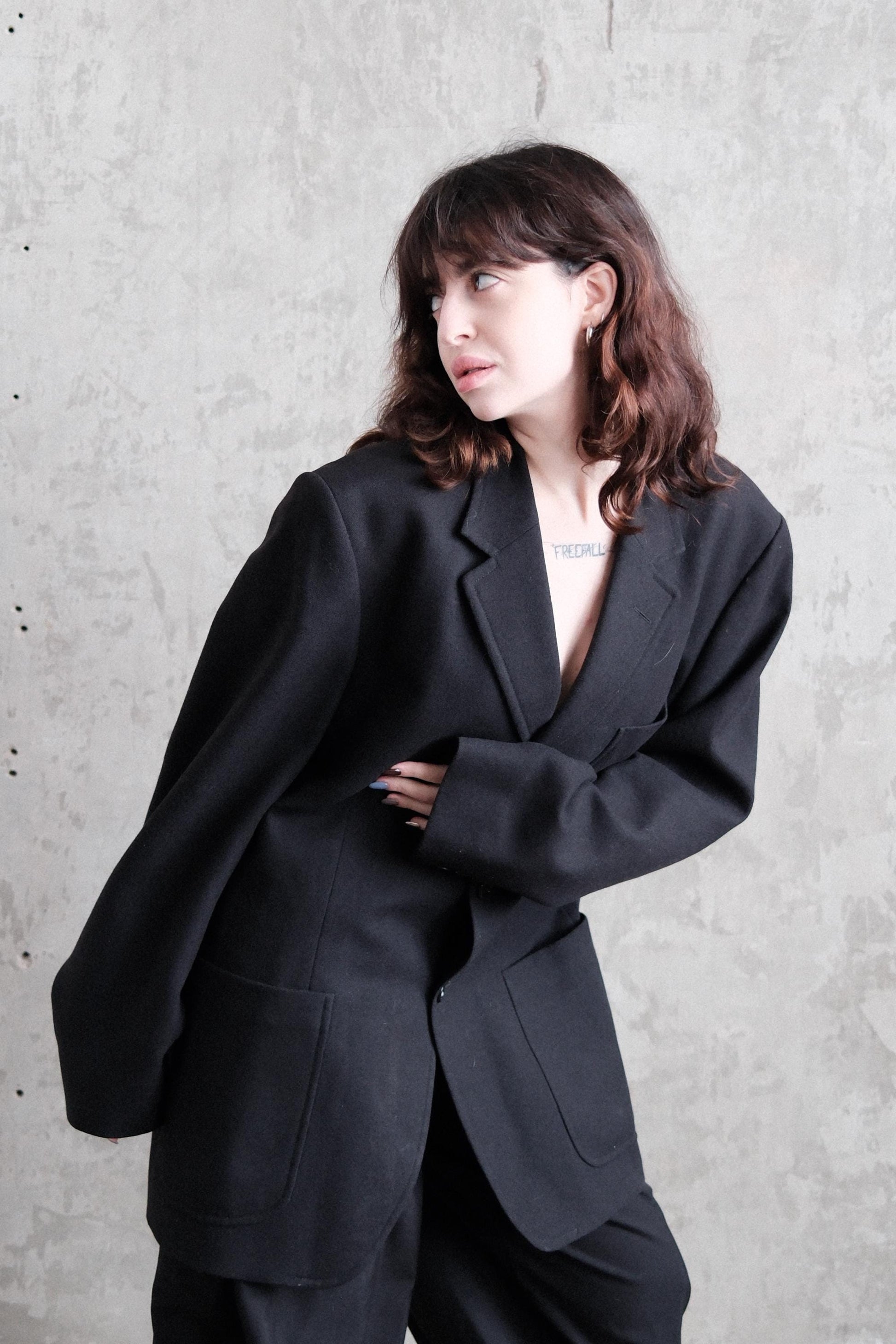 A woman in a black suit is standing against a concrete wall, looking off to the side with her hands crossed.