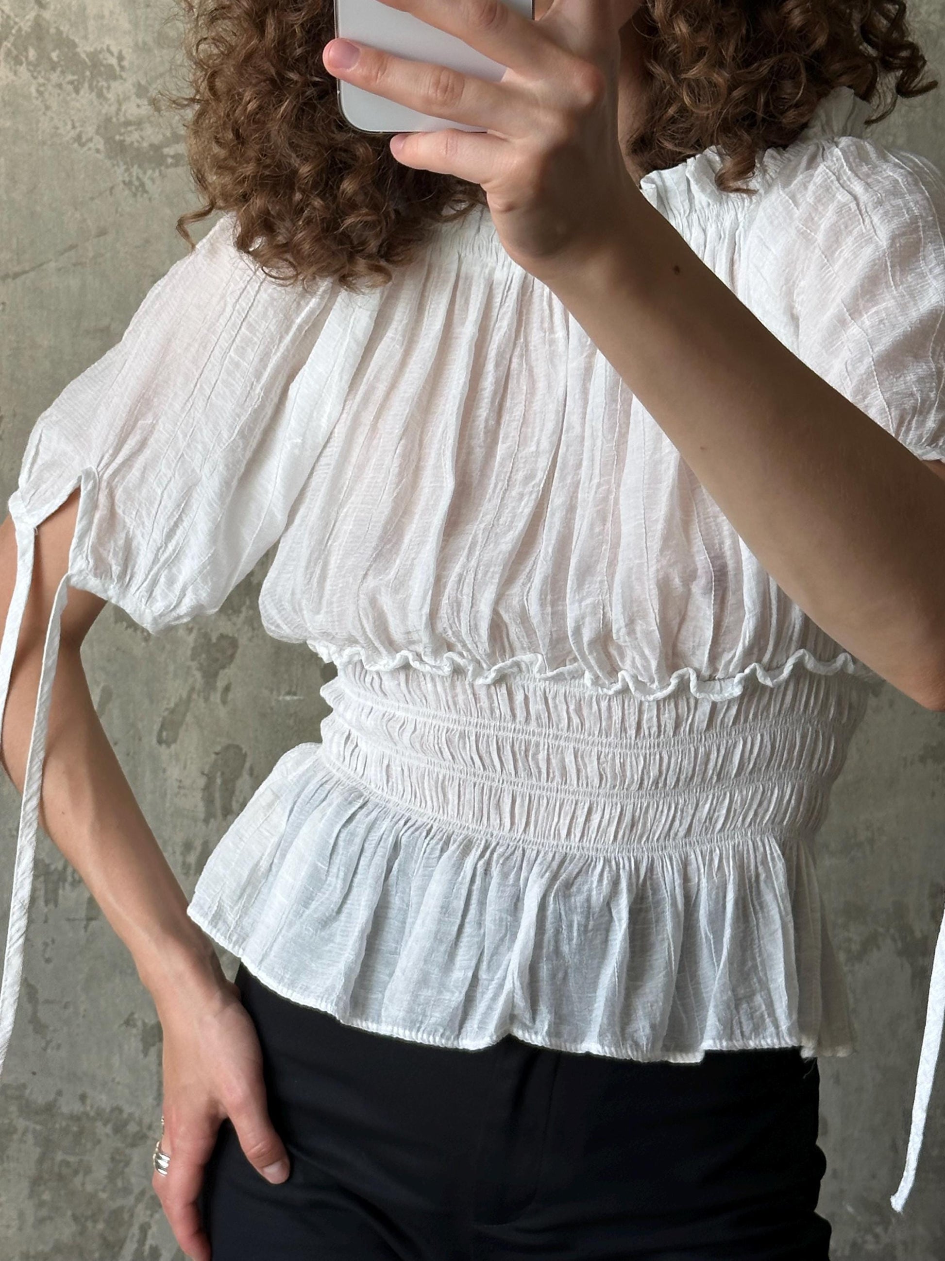 A person is taking a selfie with a white blouse featuring a ruffled collar and a white ruffled skirt.