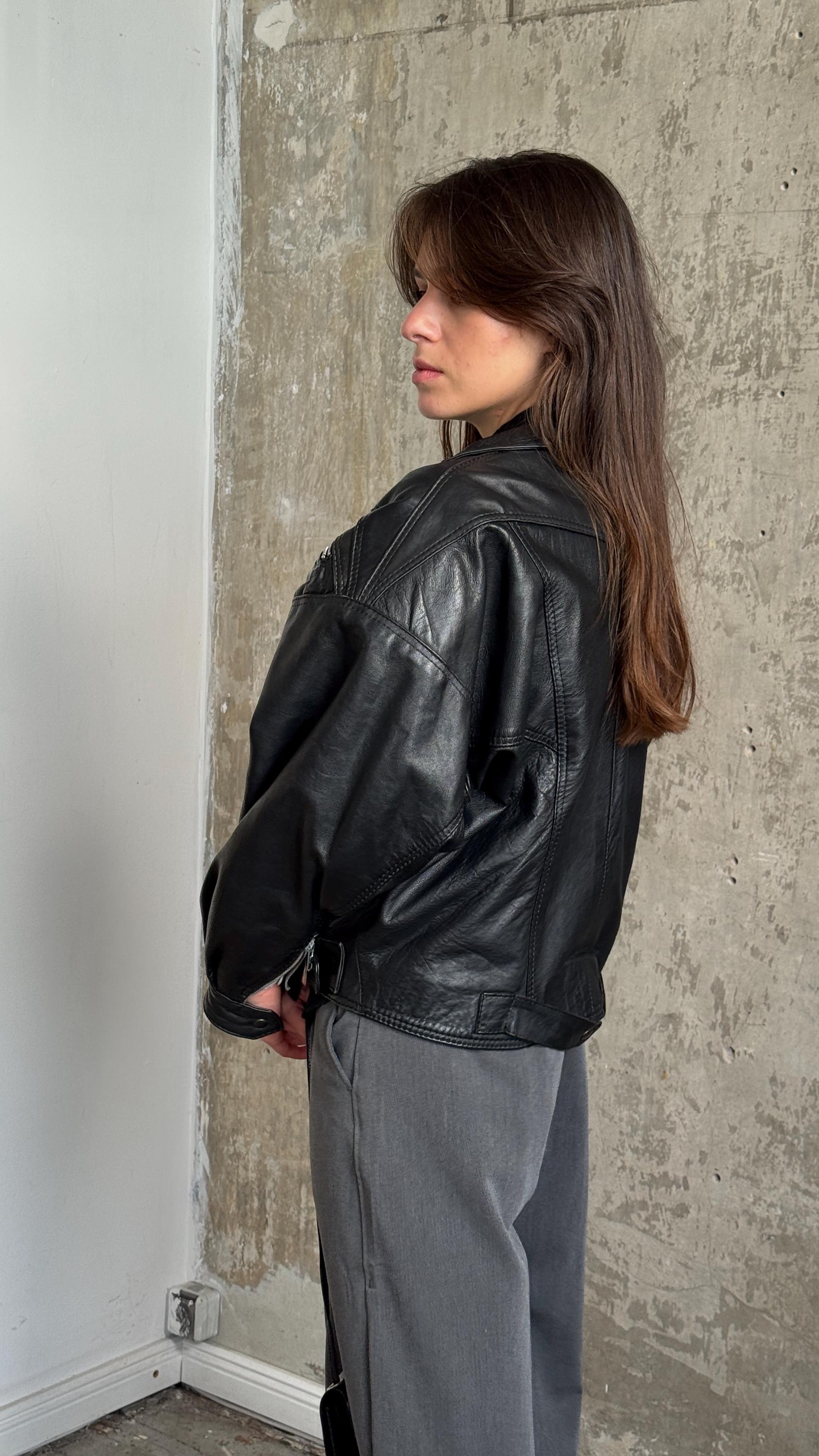 A woman wearing a black leather jacket stands in front of a concrete wall, looking away from the camera.