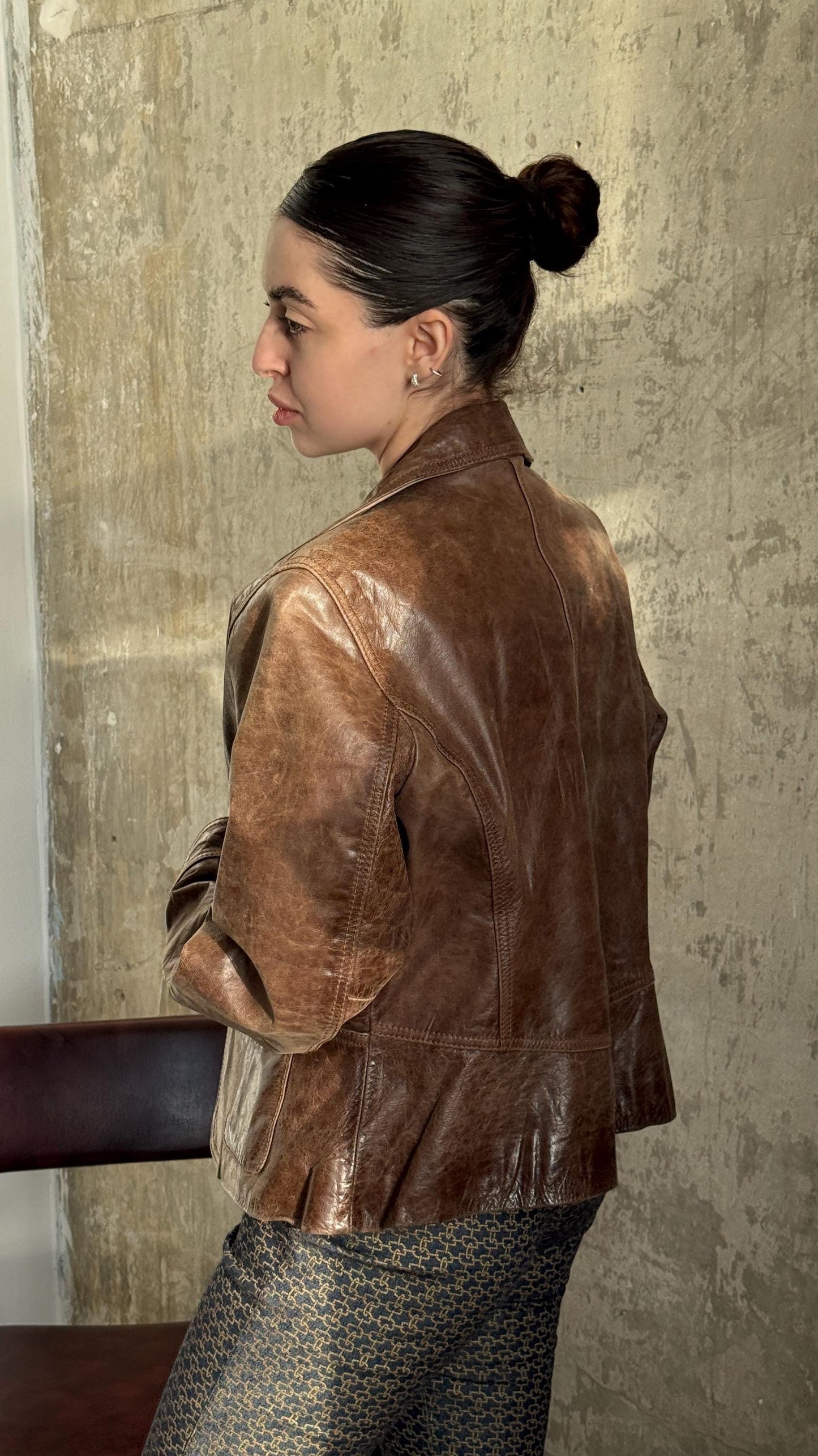 A woman wearing a brown leather jacket stands in front of a concrete wall, her back turned to the camera.