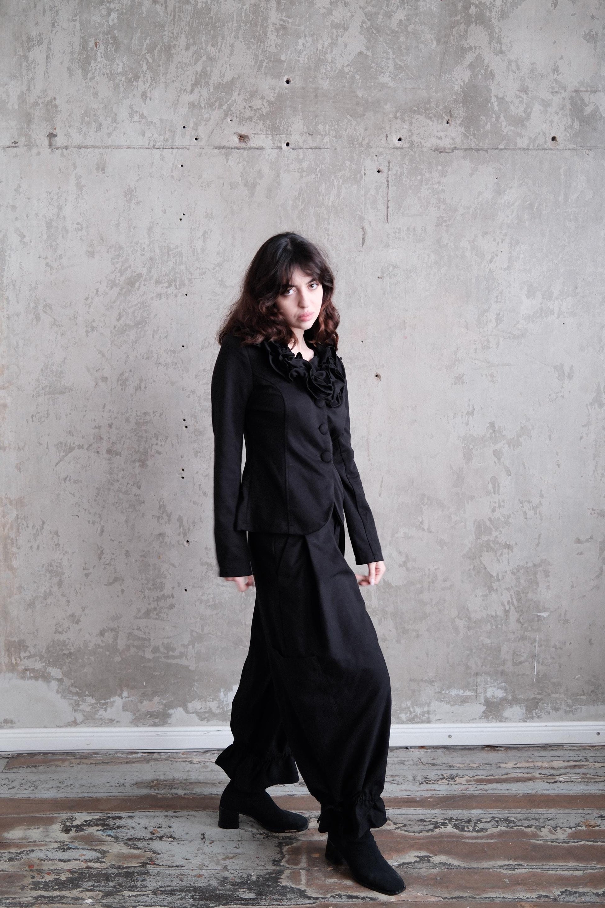 A woman in a black outfit stands in a room with a concrete wall.
