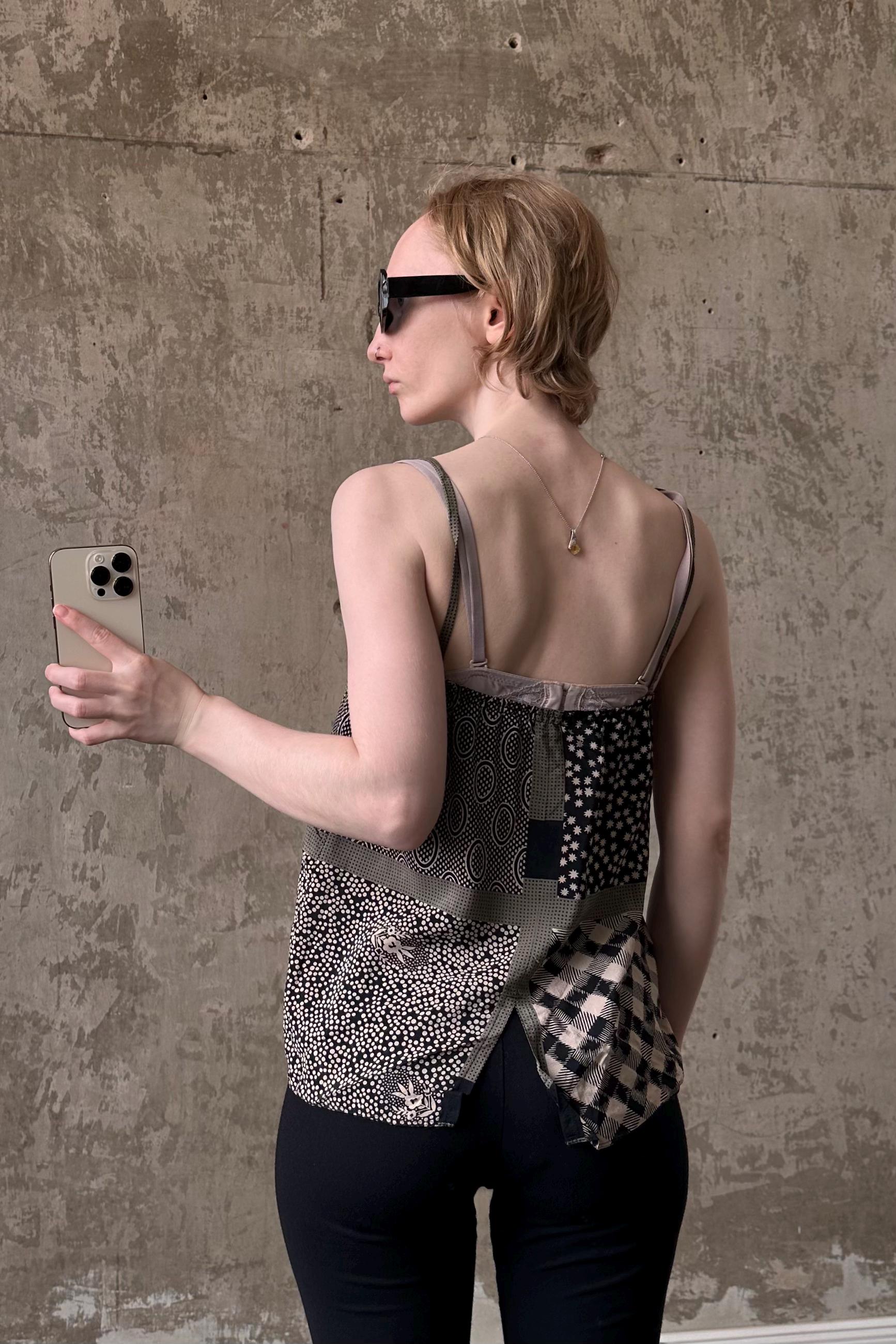 A woman wearing a patterned top and sunglasses is holding a smartphone and looking at it, with a neutral expression on her face.