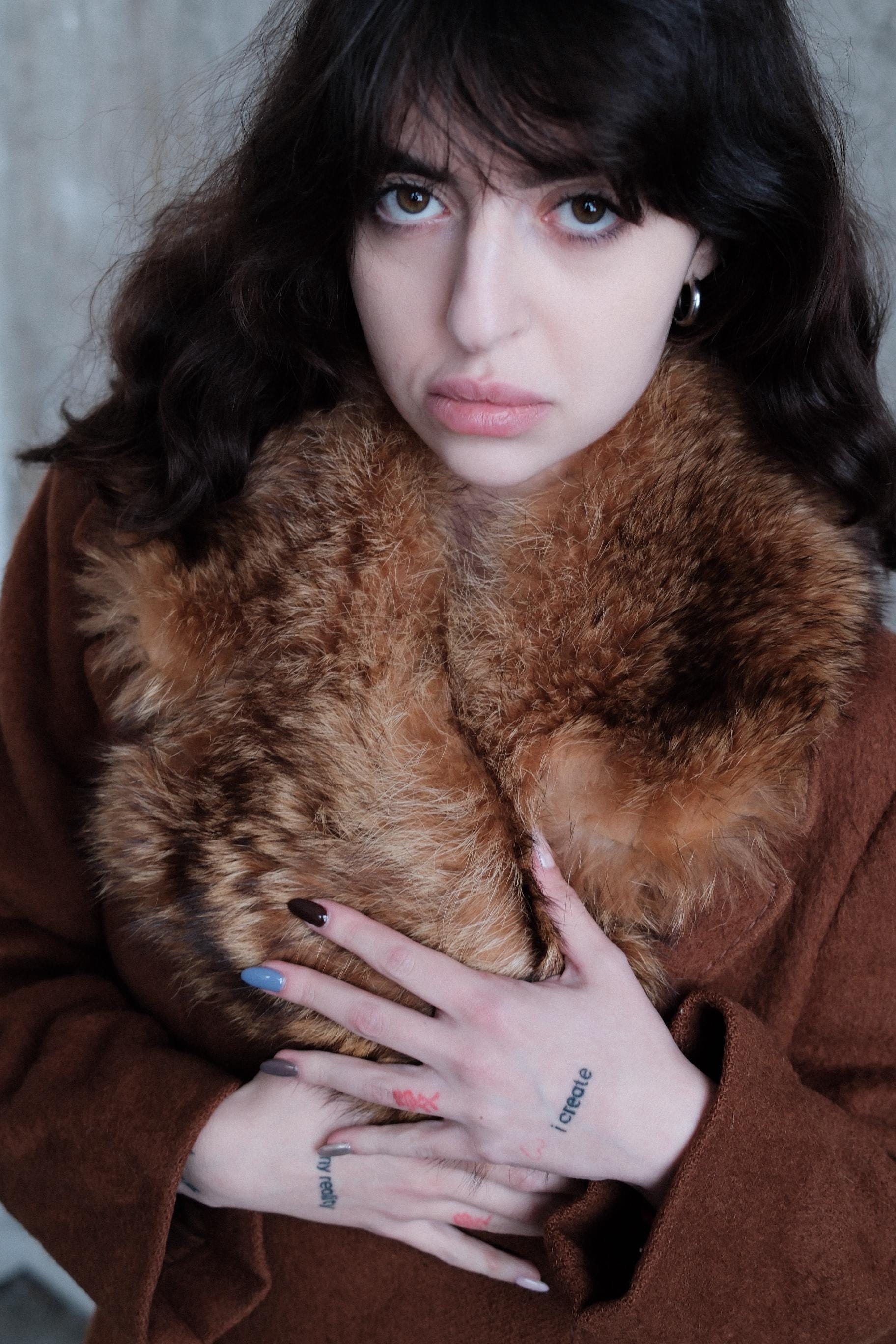 A woman with dark hair and tattoos is lying on a brown fur coat, looking up at the camera with a serious expression.