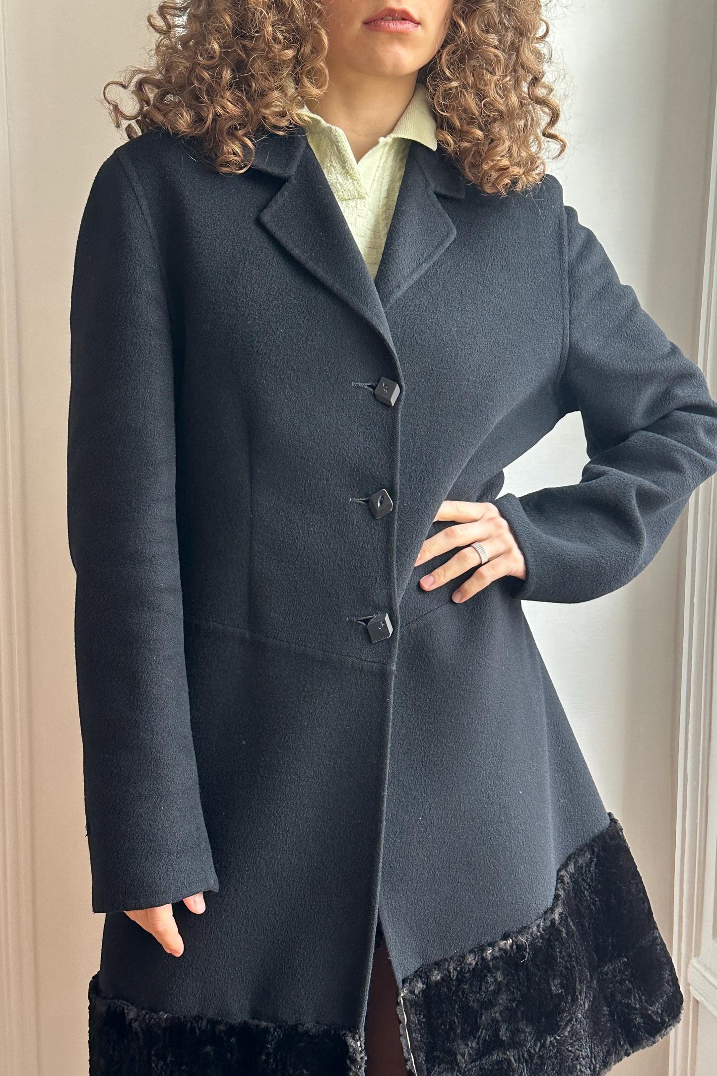 A woman wearing a dark gray coat with a fur trim, standing in front of a window.