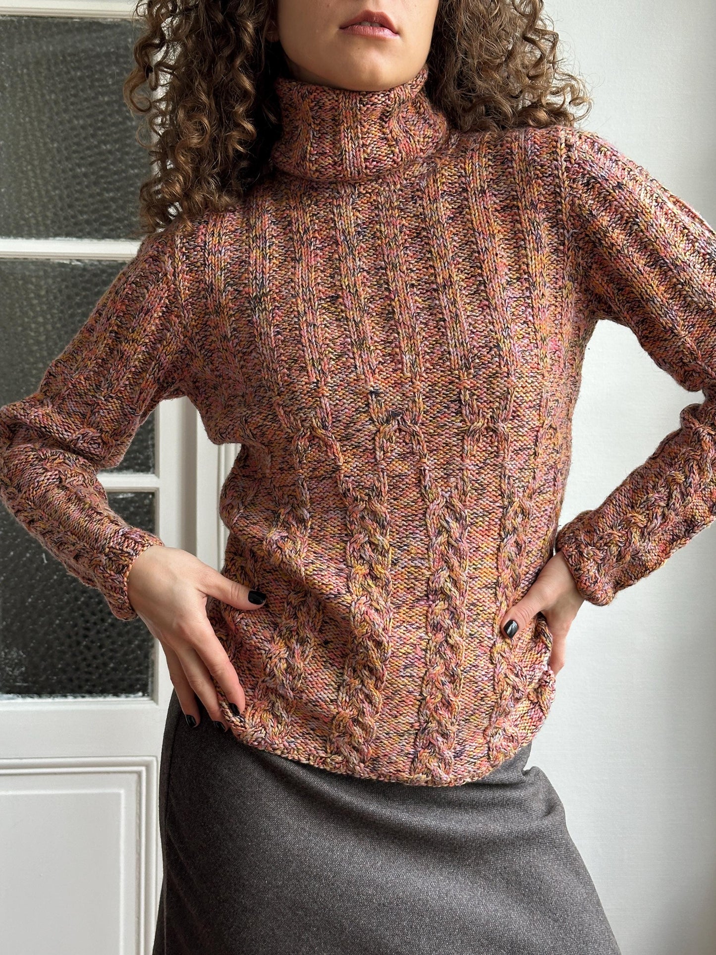 A person wearing a multicolored knitted sweater is posing with their hands on their hips, against a plain white background.
