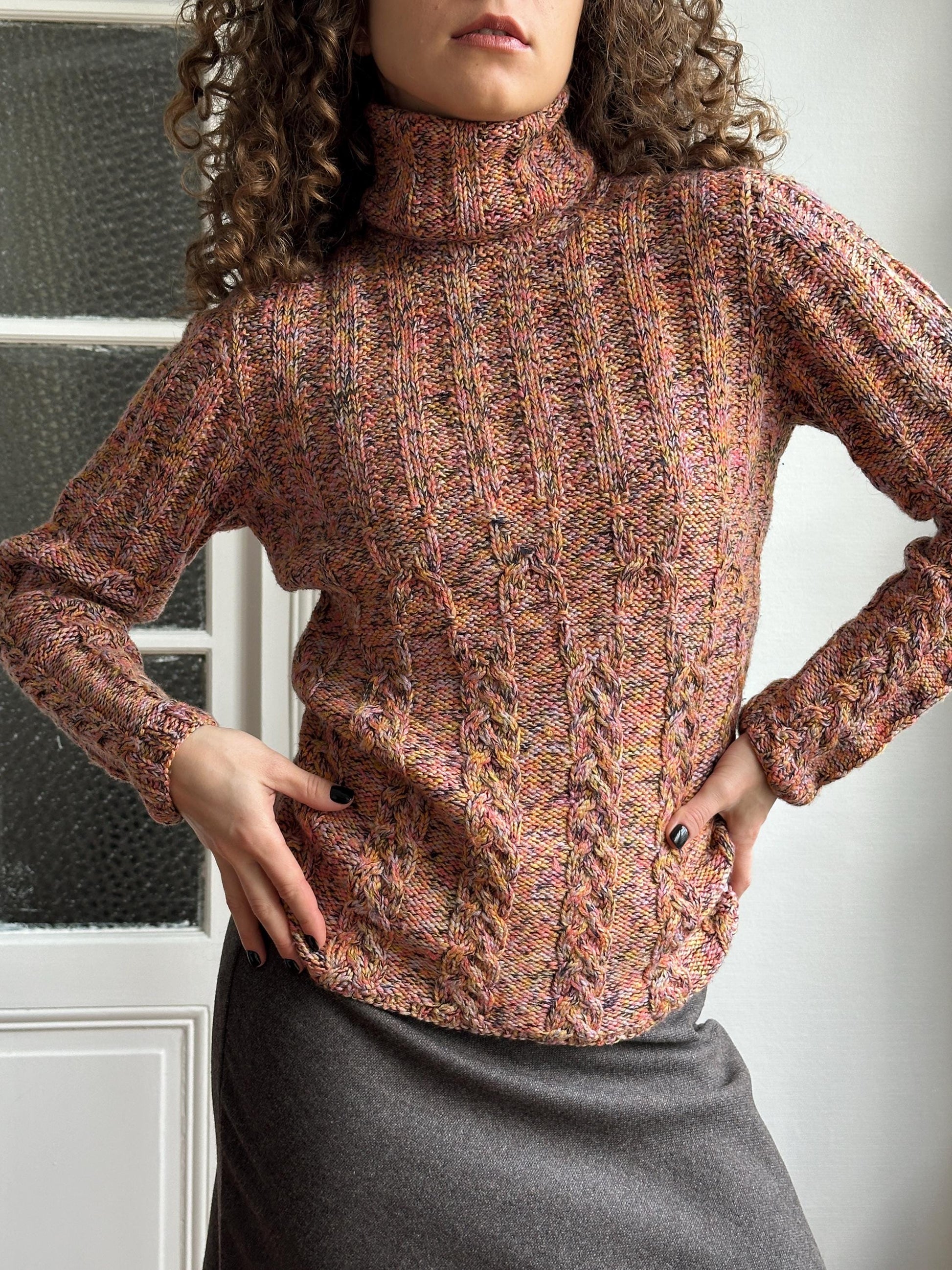 A person wearing a multicolored knitted sweater is posing with their hands on their hips, against a plain white background.